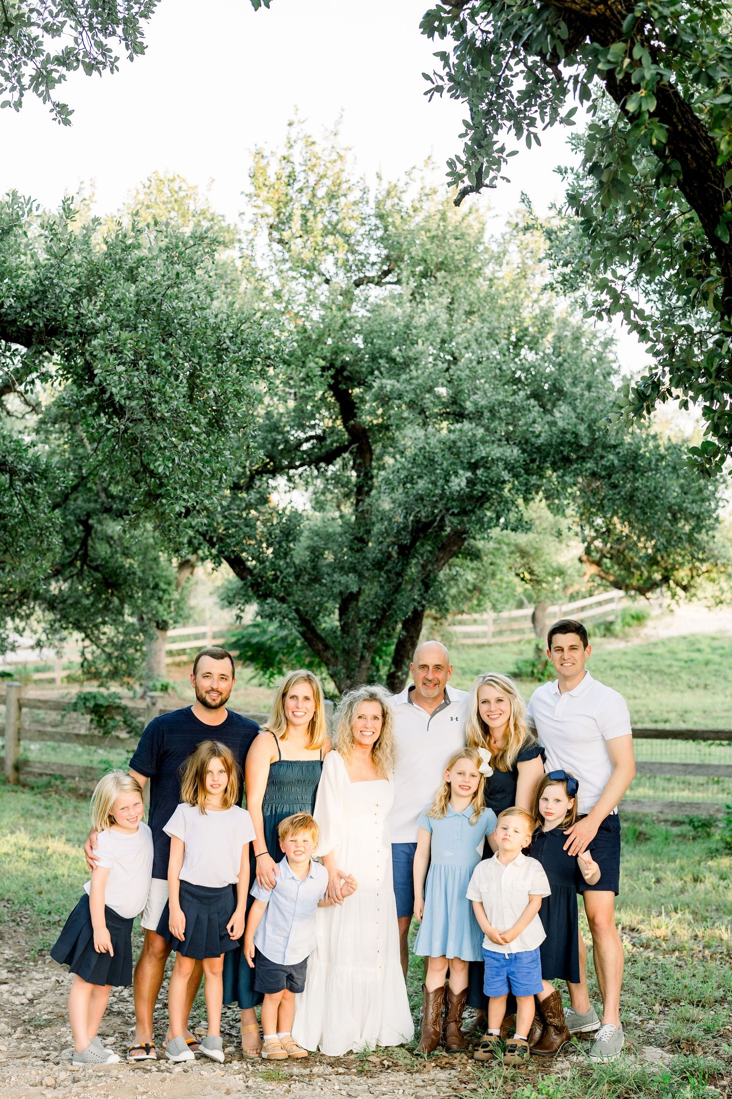 Large family portrait outdoors under a tree, adults and children in coordinated navy and white outfits on a sunny day.