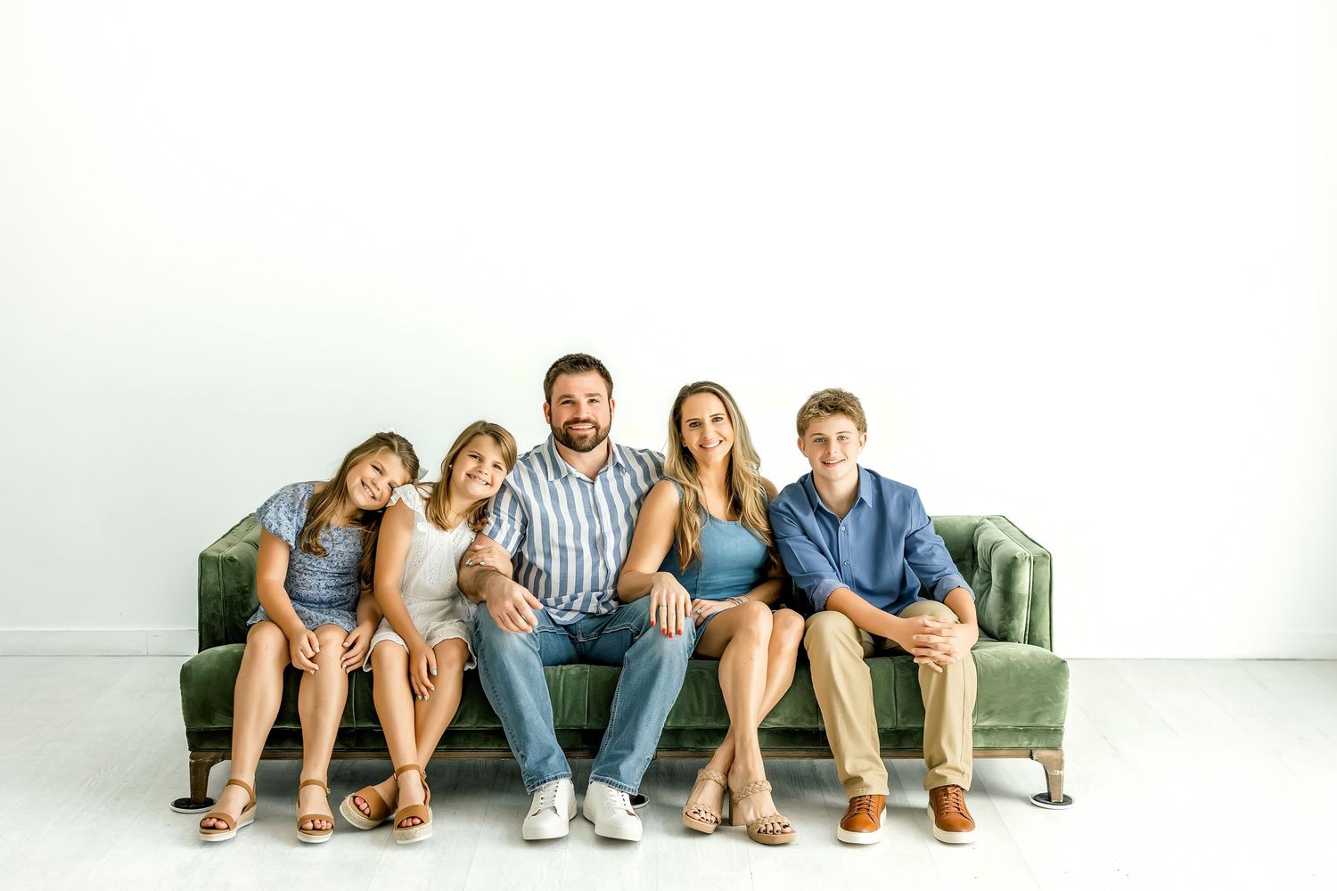 Family of five sitting closely together on a green velvet couch in a bright white studio.