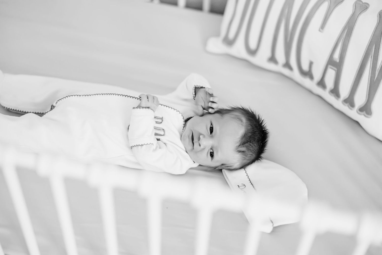 Black and white photo of newborn baby boy named Duncan lying in white crib, smiling softly at camera.