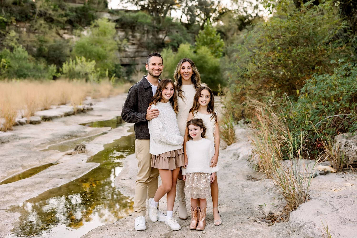 Family of five posing by rocky creek surrounded by lush greenery in neutral outfits.