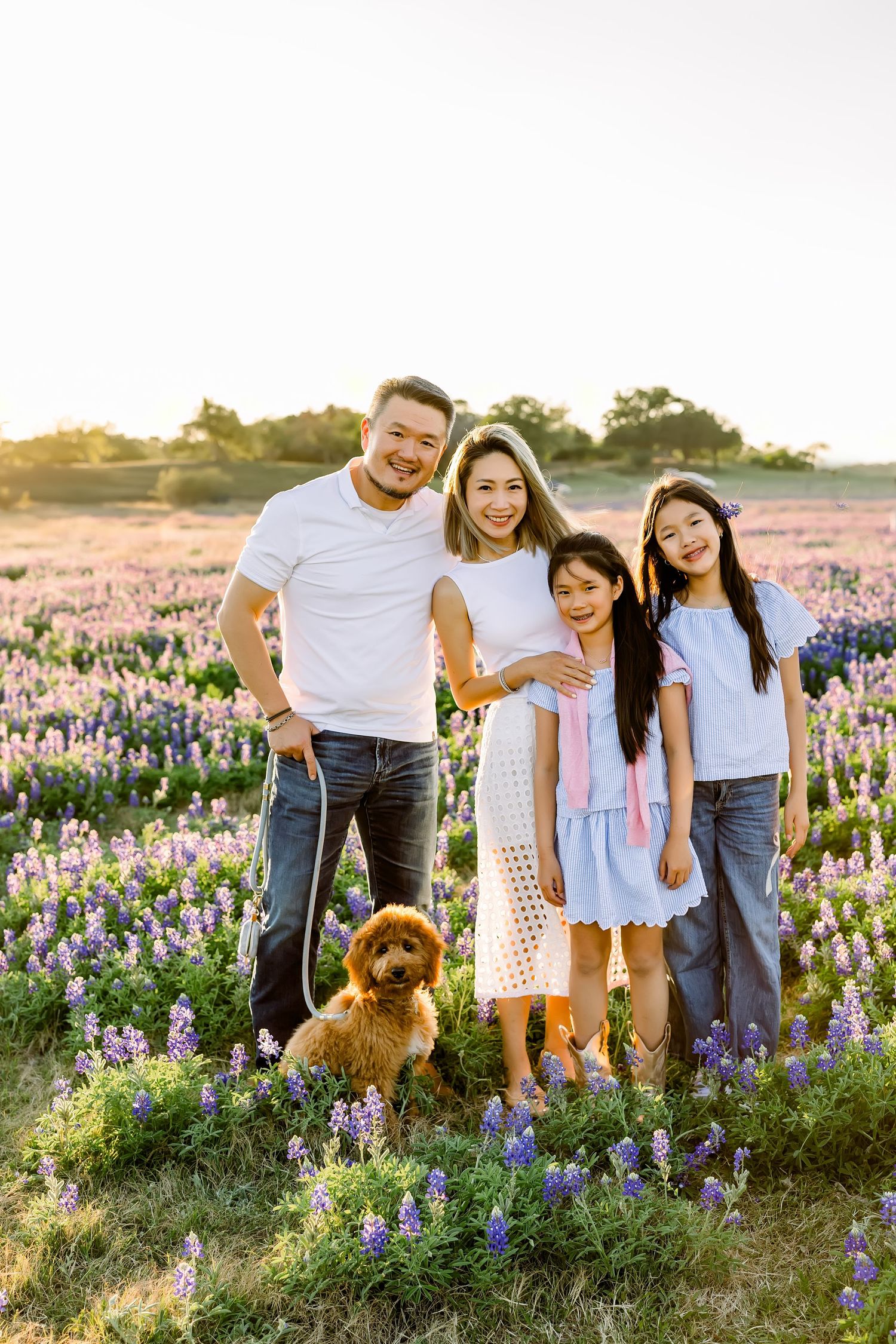 Smiling Asian family with dog posing in purple wildflower field at sunset.