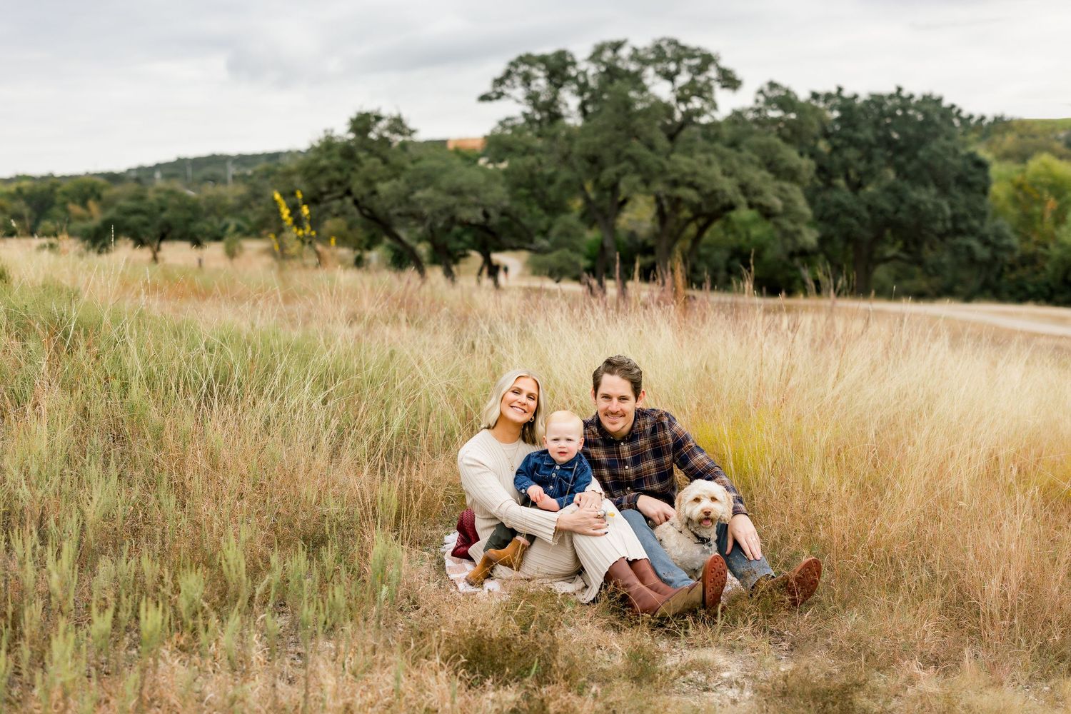 Family of three sits together in a golden field with their yellow Labrador on a cloudy morning mini session at Bee Cave Park in BeeCave, TX.