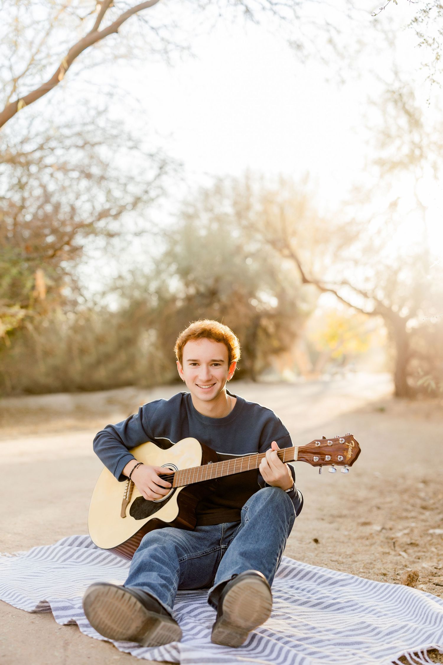 Smiling teenage boy playing acoustic guitar on a blanket outdoors in warm sunlight.