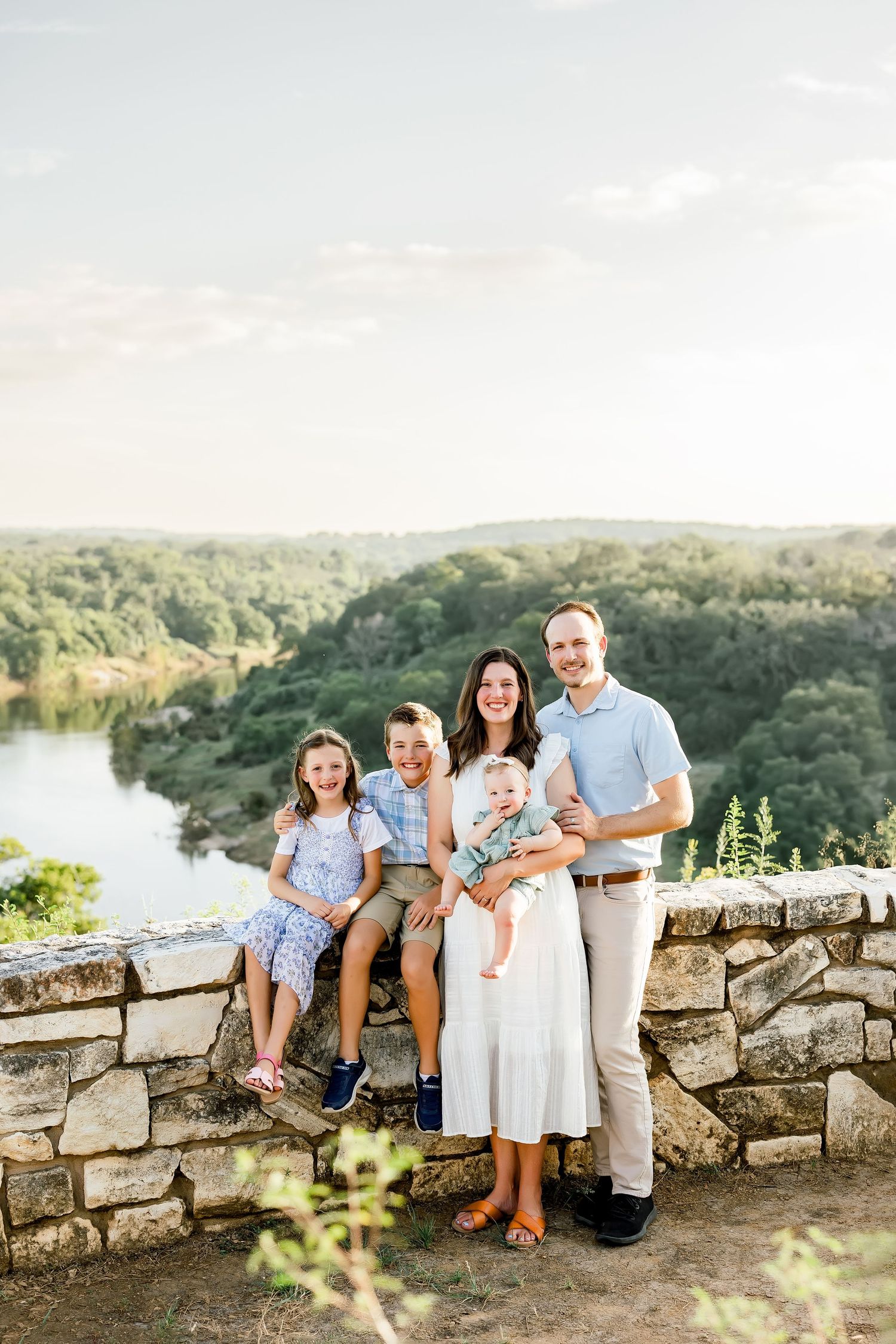 Family of five posing on stone wall overlook with river and lush green trees in background on sunny day.