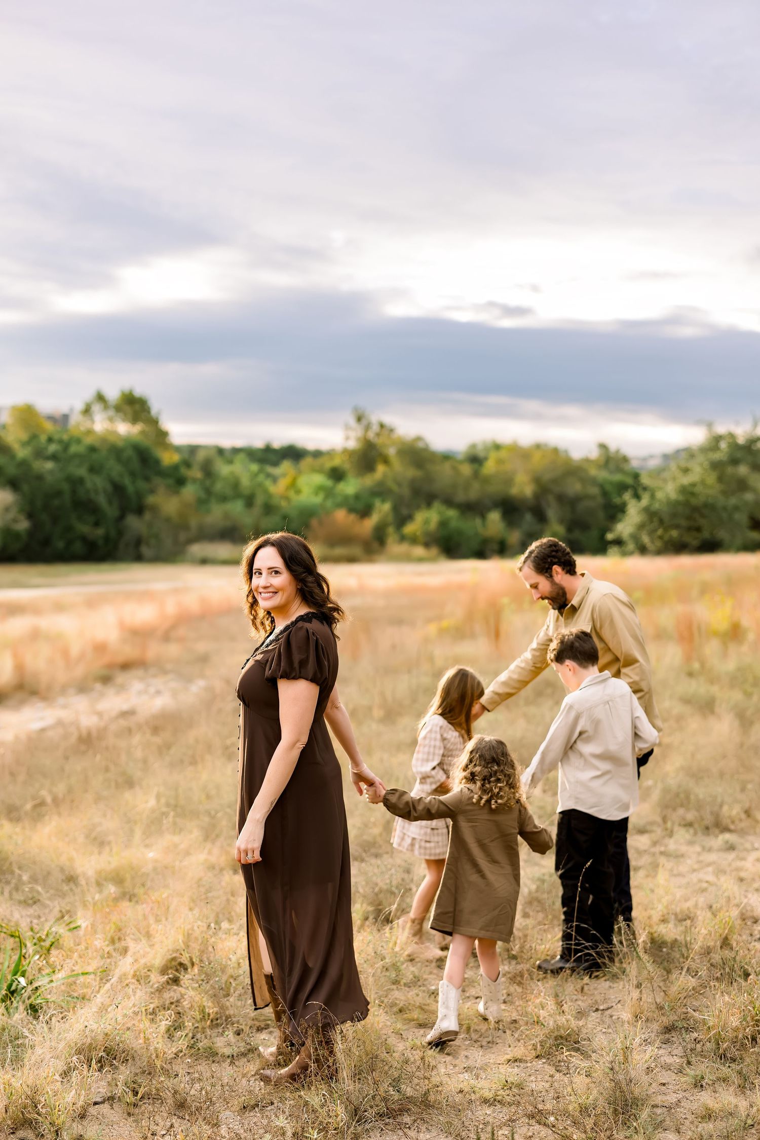 Family of four holding hands in golden field at sunset, mother in brown dress smiling back at camera.
