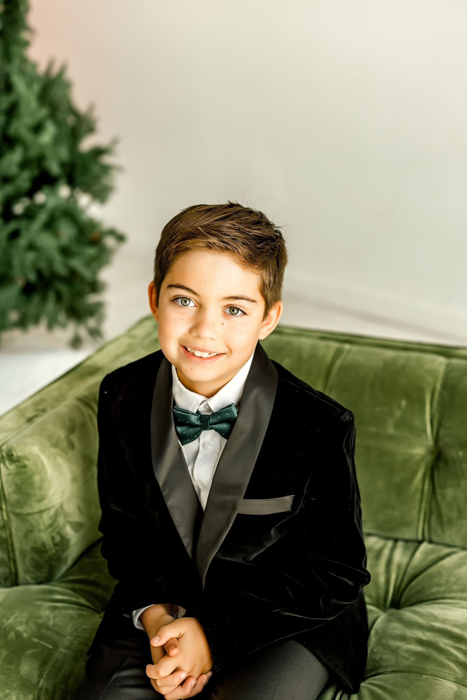 Smiling boy in black velvet tuxedo with green bow tie seated on green velvet sofa near Christmas tree.