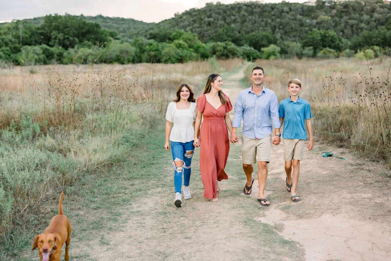 Family of four walking a dog on a dirt path through an open field at a mini session in Commons Ford Ranch in Bee Cave, Tx.