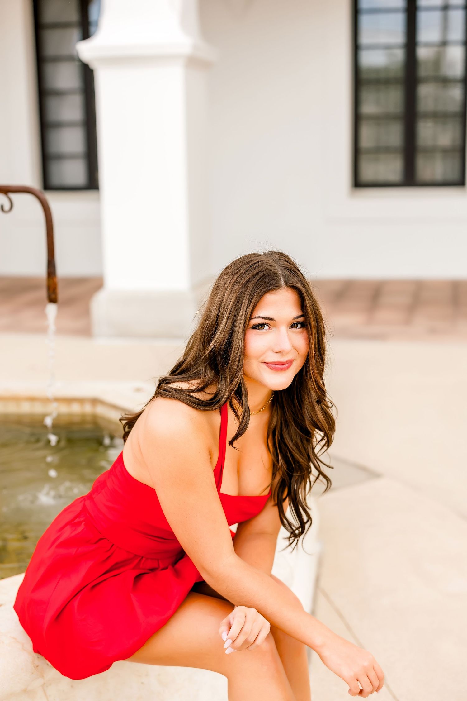 Young woman in red dress sitting by fountain smiling at camera near white building.