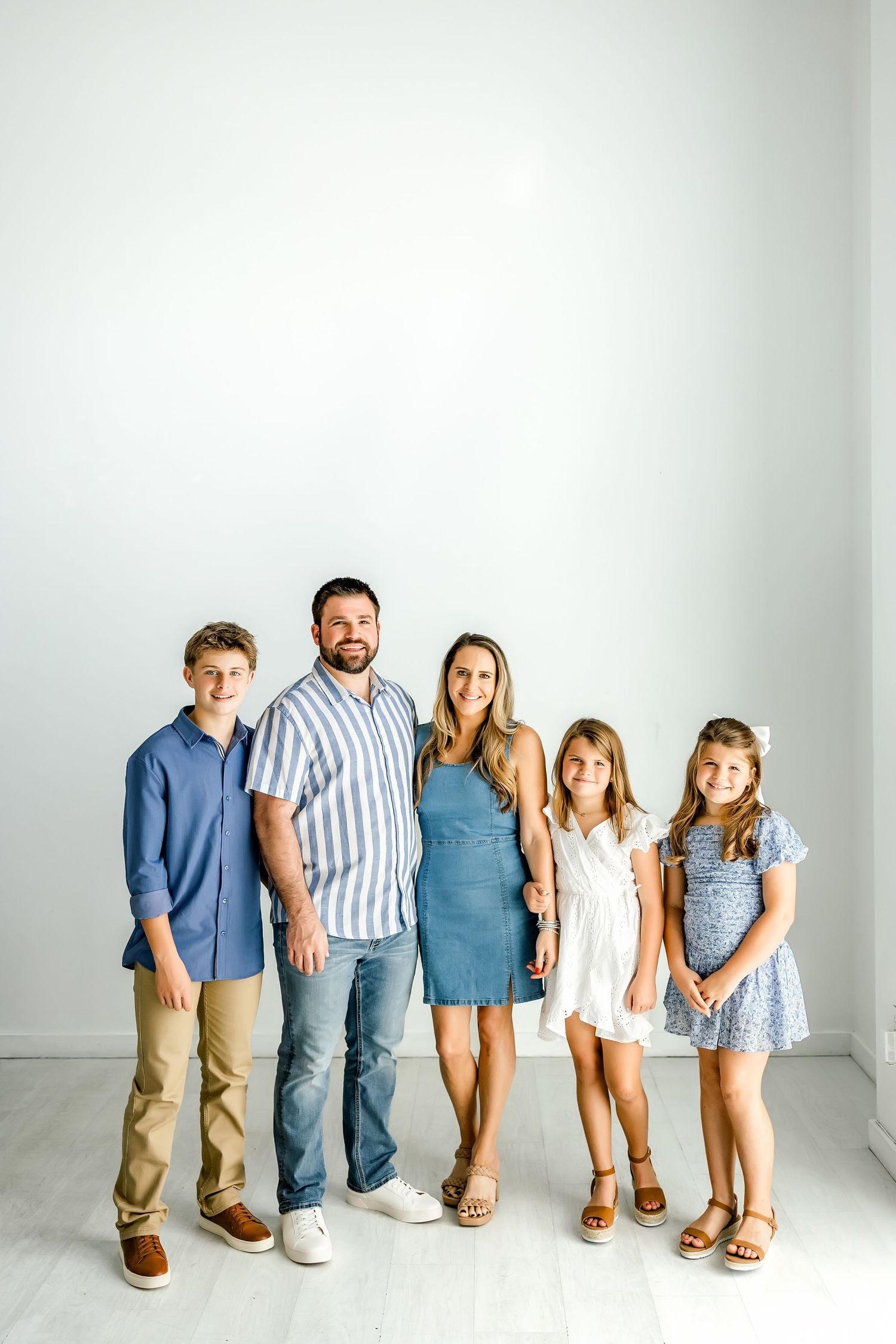 Family of five smiling together in white studio, parents with three children in coordinated blue and white outfits.
