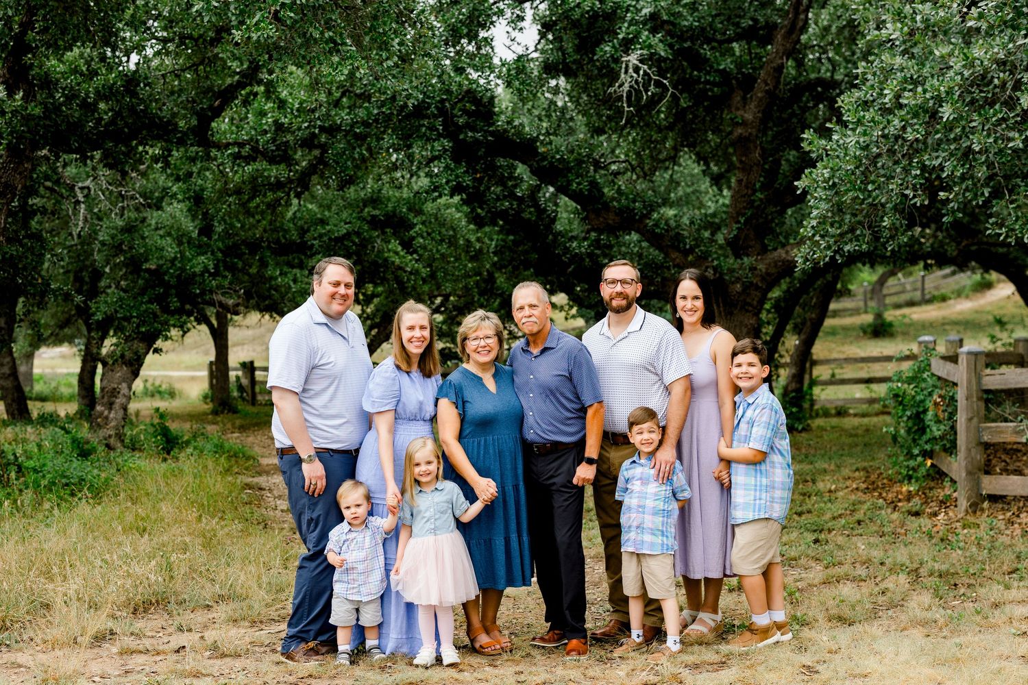 Family portrait of multigenerational group with children posing outdoors under oak trees in a green park setting.