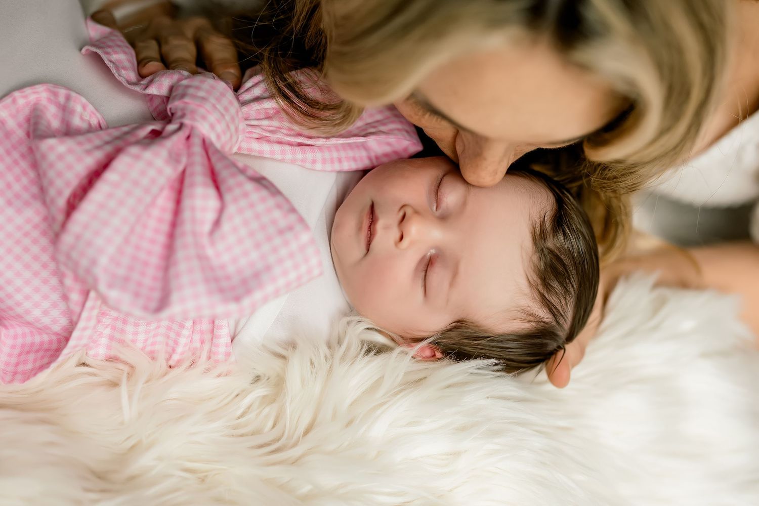 A mother tenderly kisses her sleeping newborn baby on a soft white fur blanket.