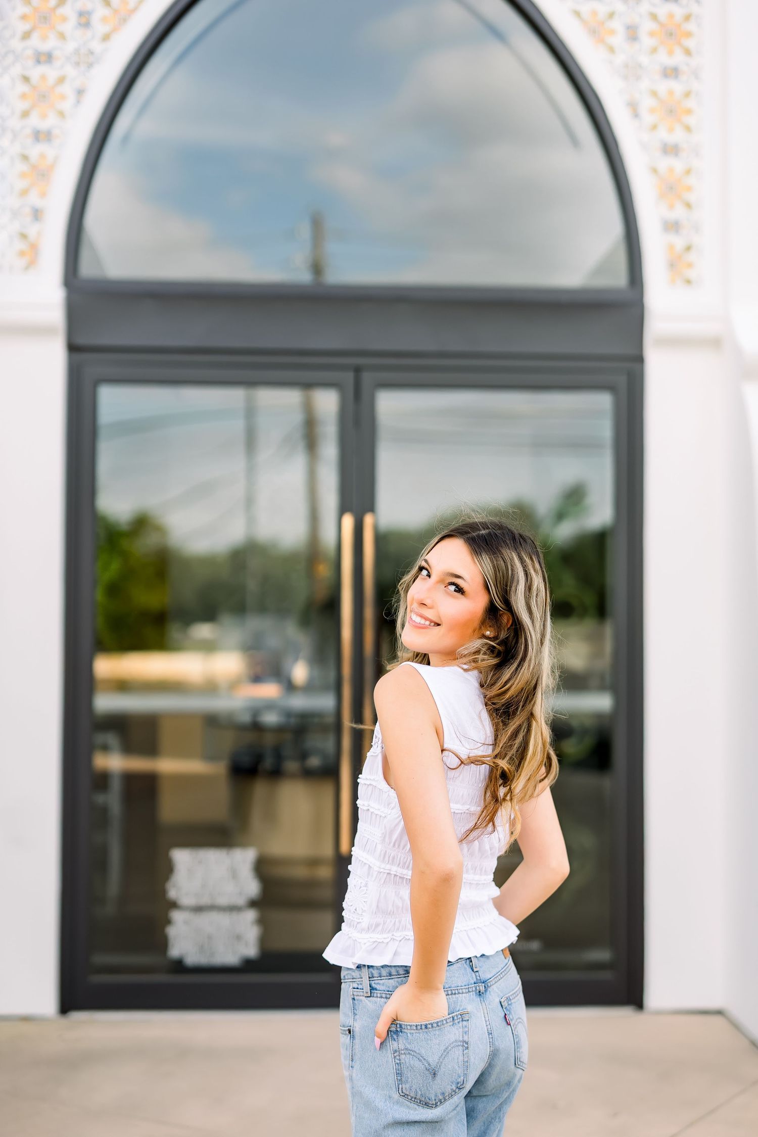 Young woman in white lace top and jeans smiling over shoulder in front of arched glass doors.
