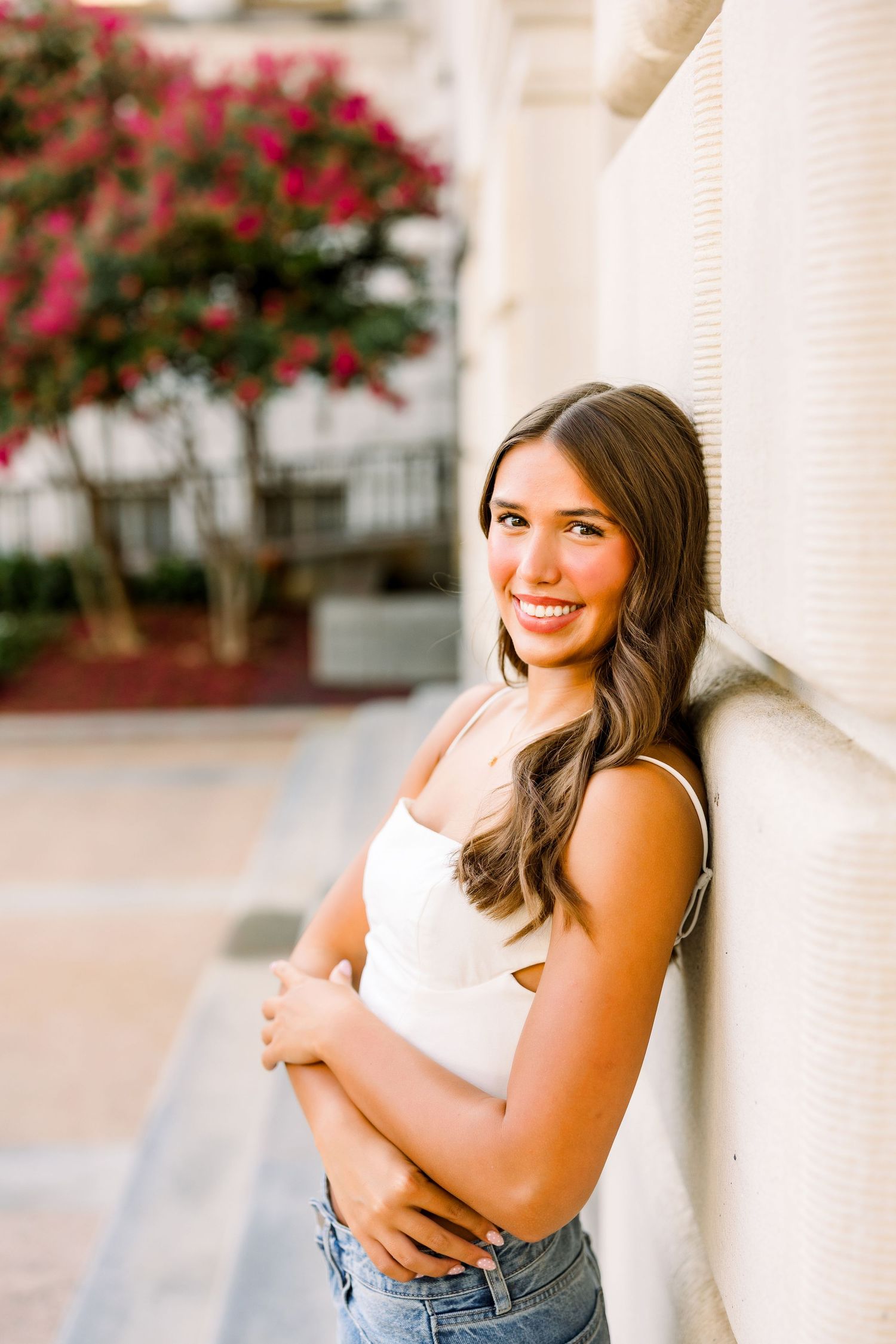 Smiling young woman in white top and jeans leaning against a wall with pink flowering trees in background.