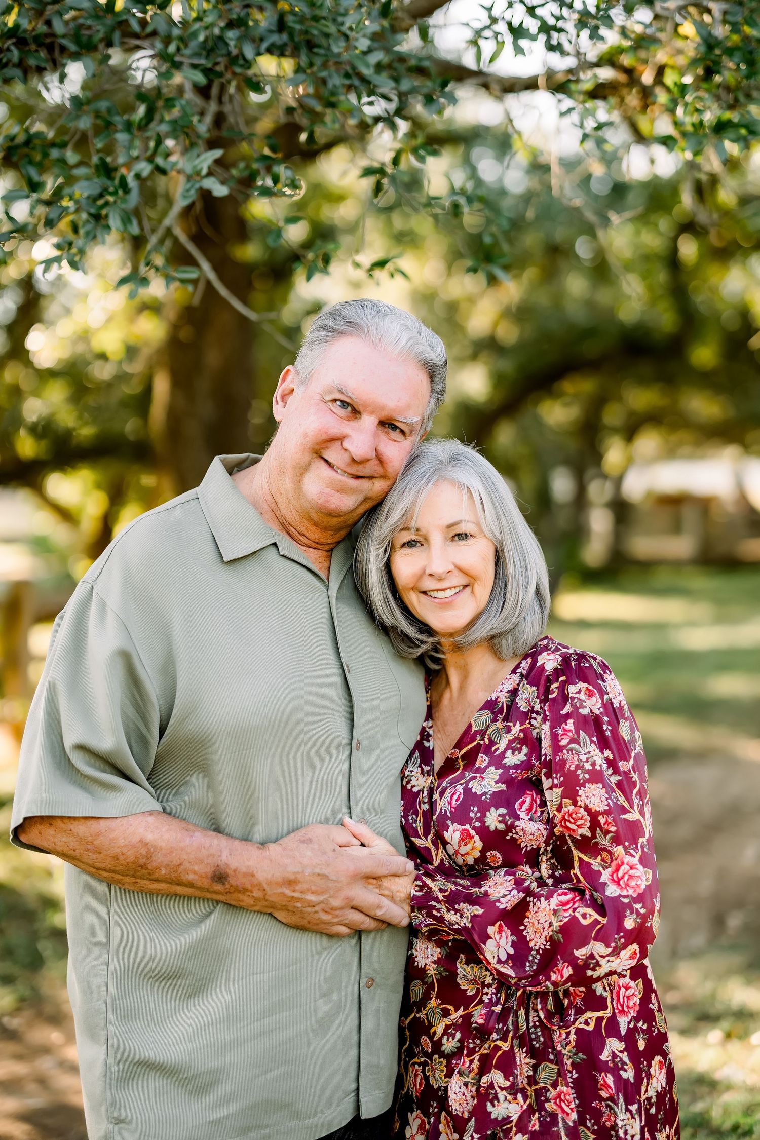 Smiling older couple embracing outdoors under green trees, woman in floral dress and man in sage shirt.