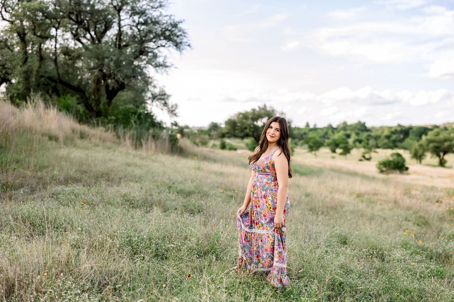 A young woman in a colorful floral maxi dress and flower crown stands in a lush green meadow.