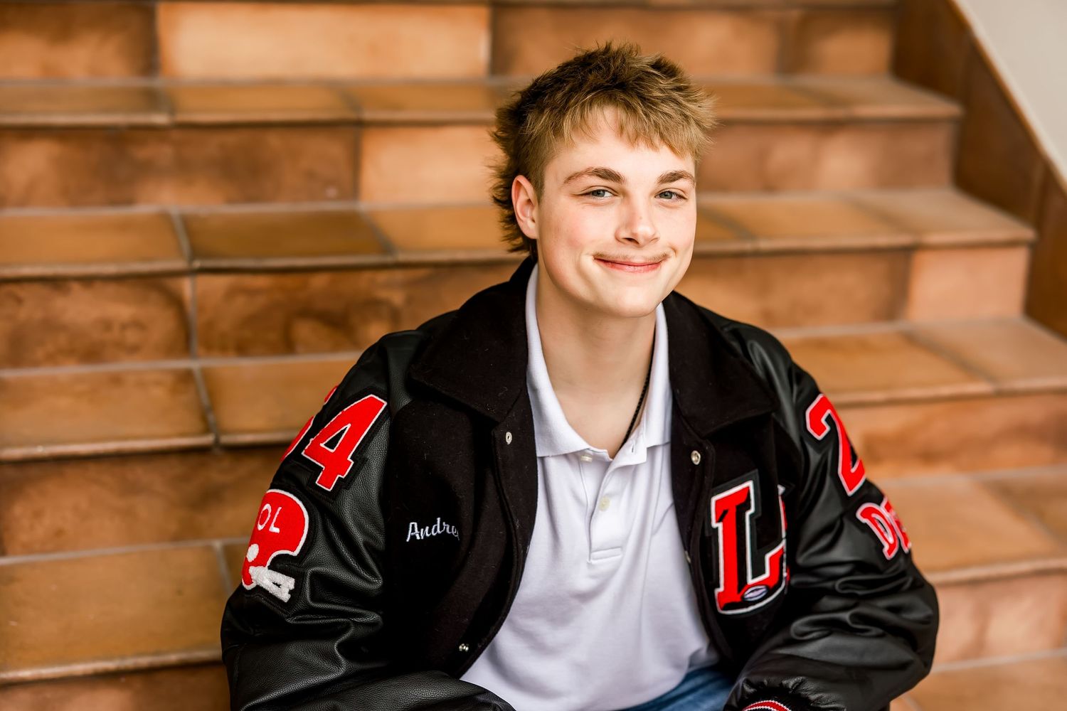 Smiling teen boy in black letterman jacket sitting on brick steps outdoors.