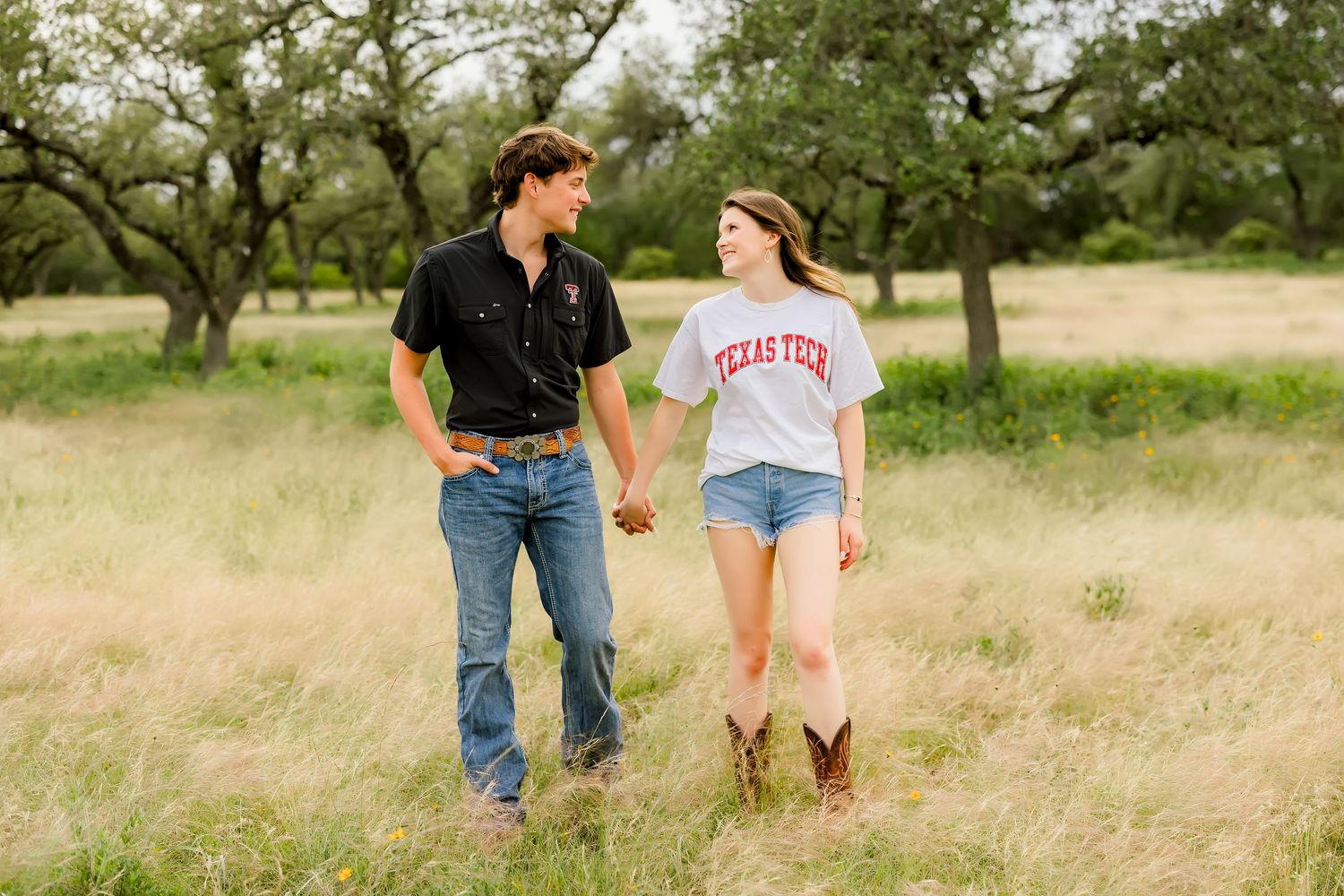 Couple holding hands walking through tall grass field with trees, woman wearing Texas Tech shirt and cowboy boots.