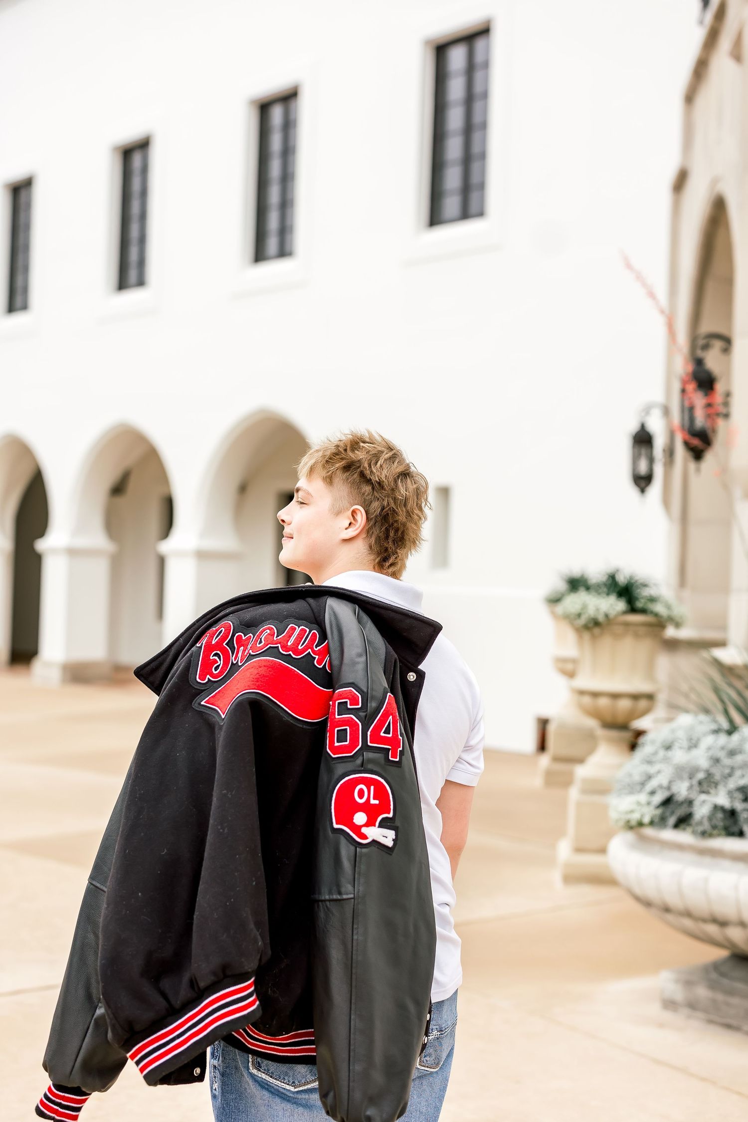 Teen boy holding black and red Brawlers letterman jacket outside white arched building.