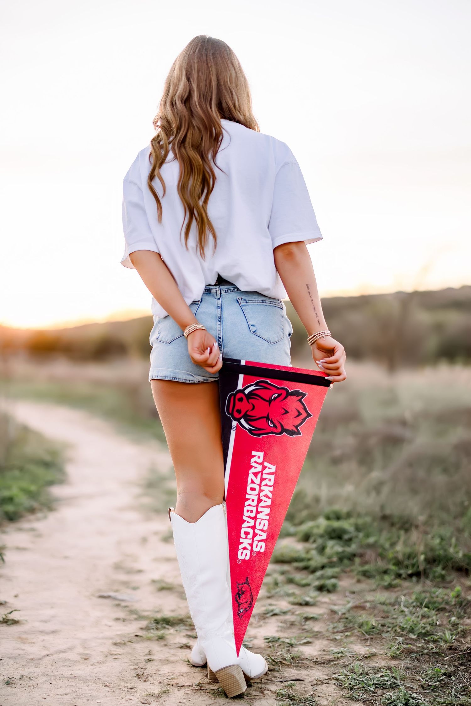 A blonde woman in denim shorts and white cowboy boots holds an Arkansas Razorbacks pennant.