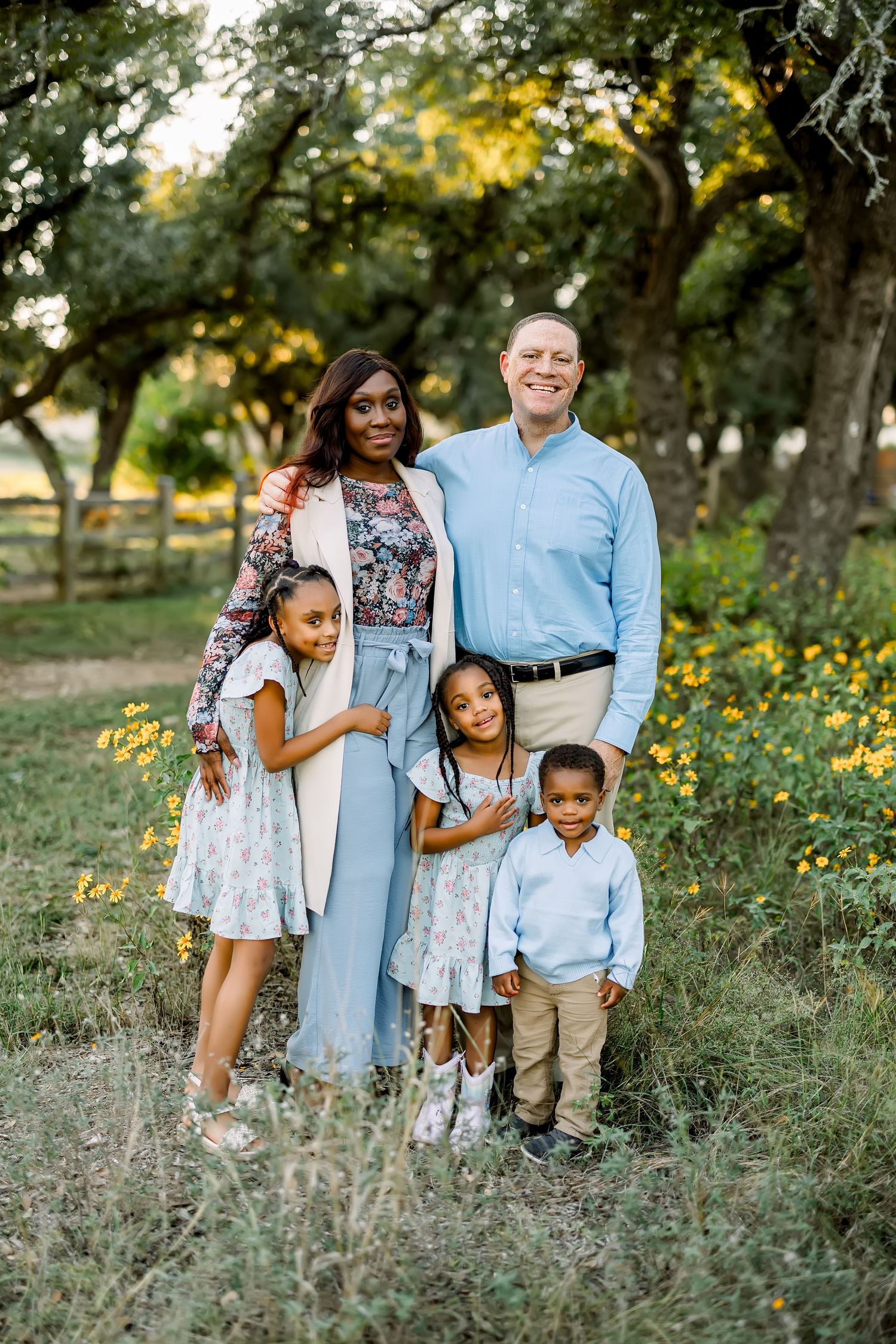 Interracial family of five posing among wildflowers under shady trees at at a mini session in Bee Cave, Tx  by Shannon Worley Photography.