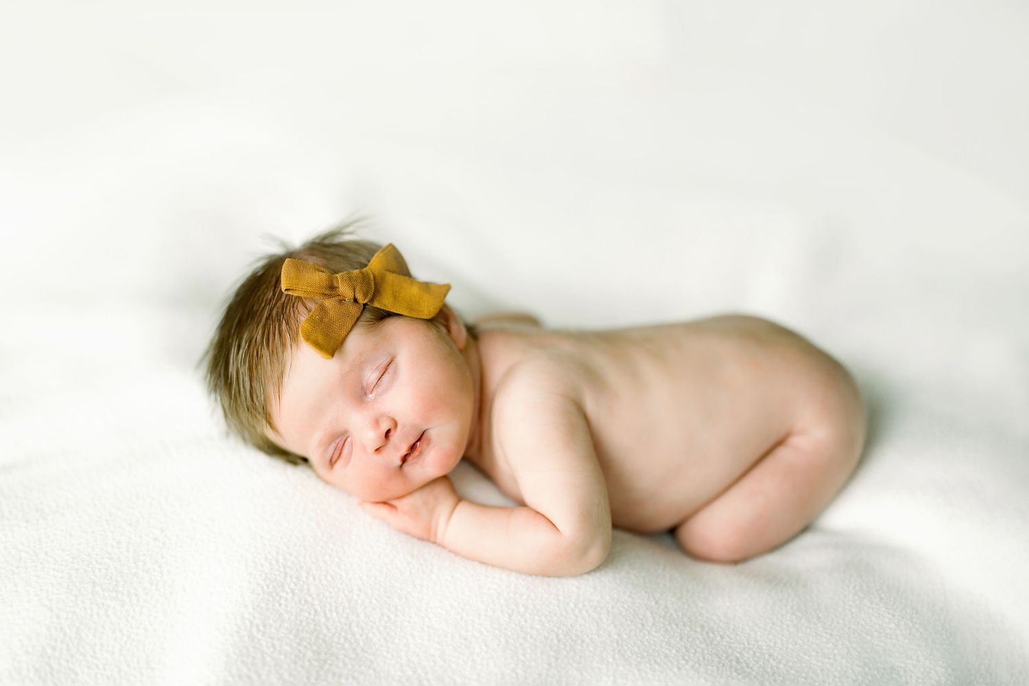 Sleeping naked newborn baby girl with a gold bow headband curled on a white blanket.