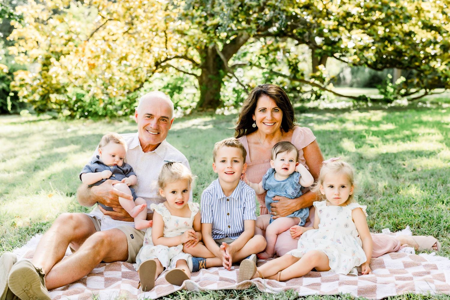 A family of grandparents and five young grandchildren smiling together on a blanket in a lush green park.