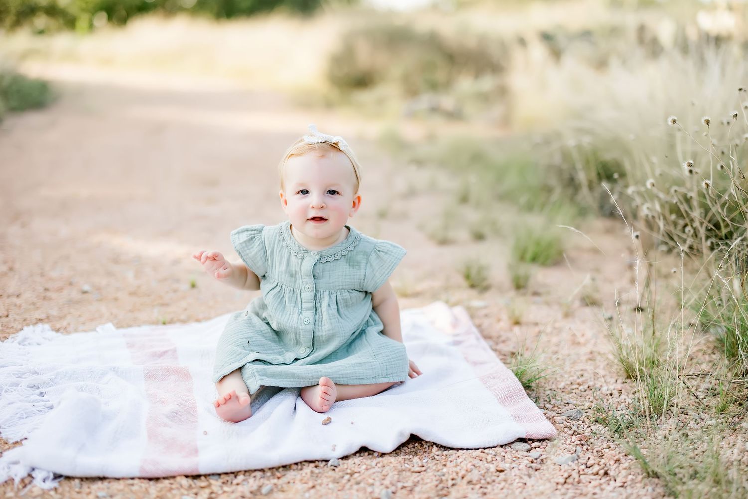 Adorable baby girl in sage green dress sitting on blanket on a sandy outdoor path.