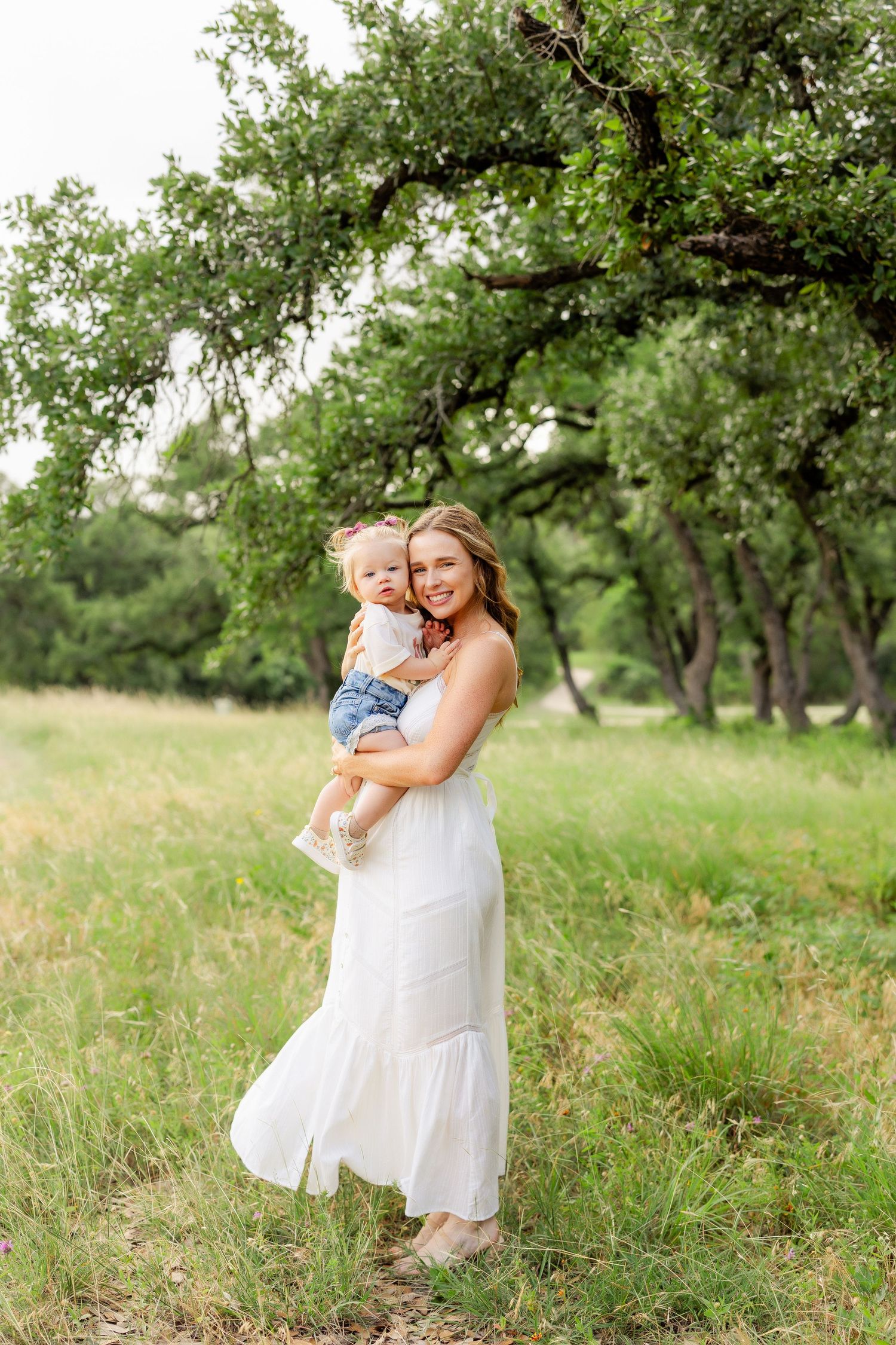 Smiling pregnant woman in white dress holding toddler daughter in lush green field.