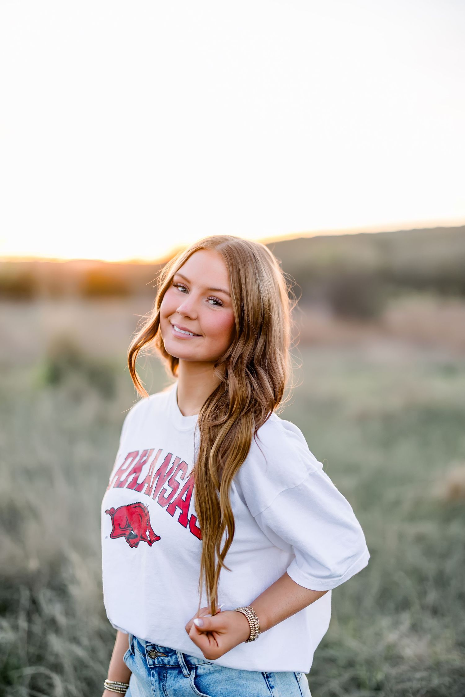 A smiling young woman wearing an Arkansas Razorbacks sweatshirt poses in a sunlit open field.