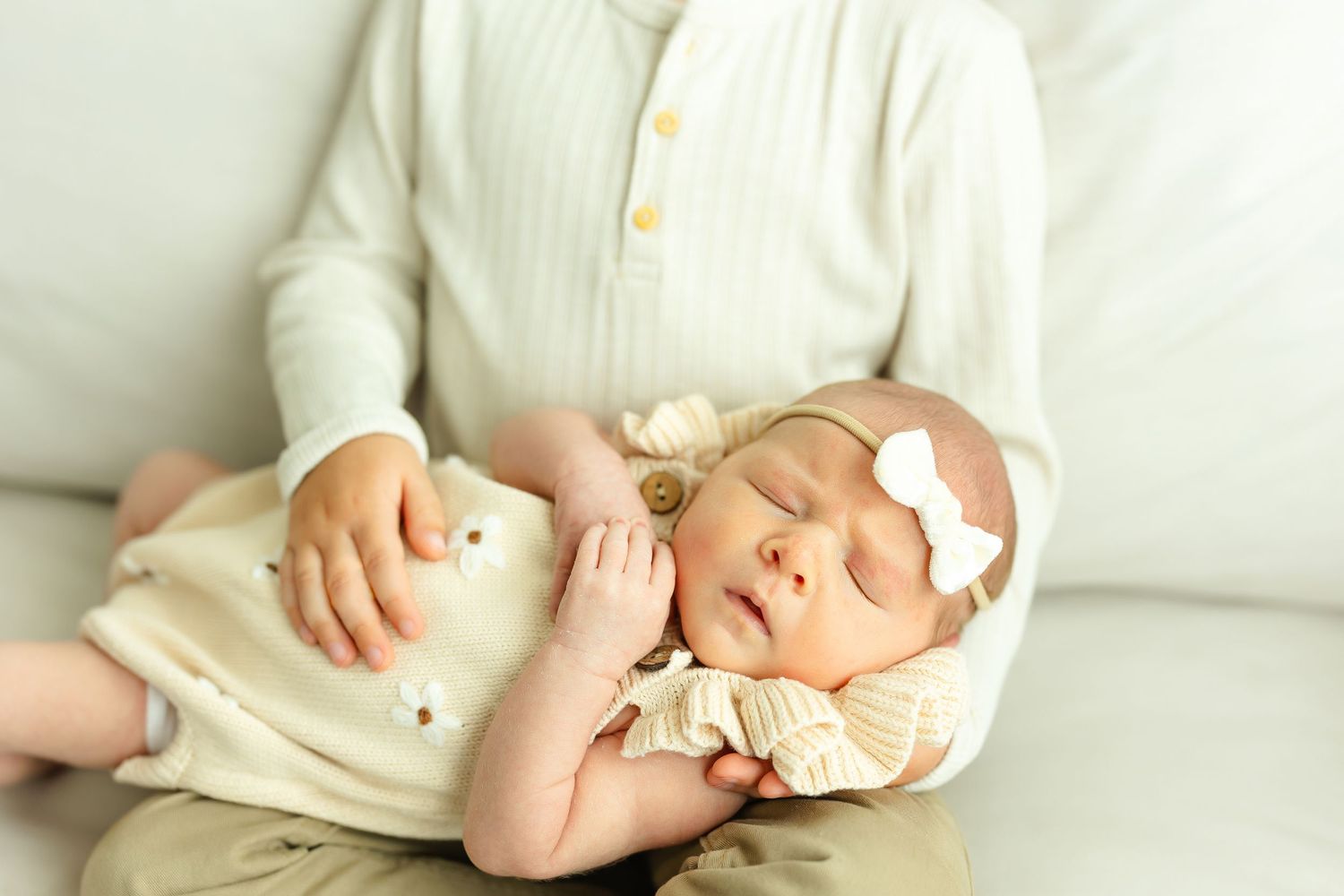 Older sibling in cream outfit gently cradling sleeping newborn baby sister with a pink bow headband.