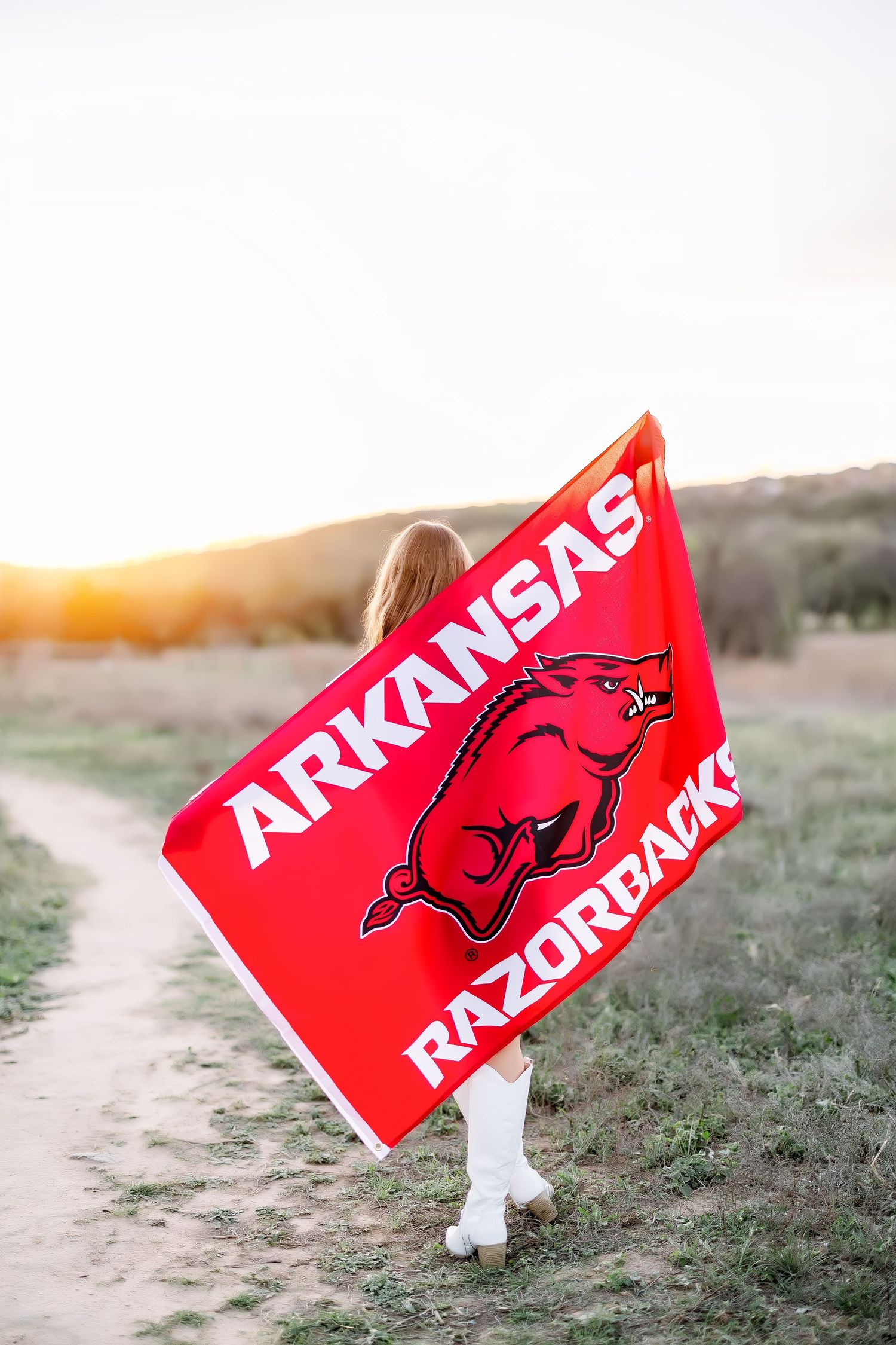 A young woman holds an Arkansas Razorbacks flag on a scenic dirt path at golden hour.