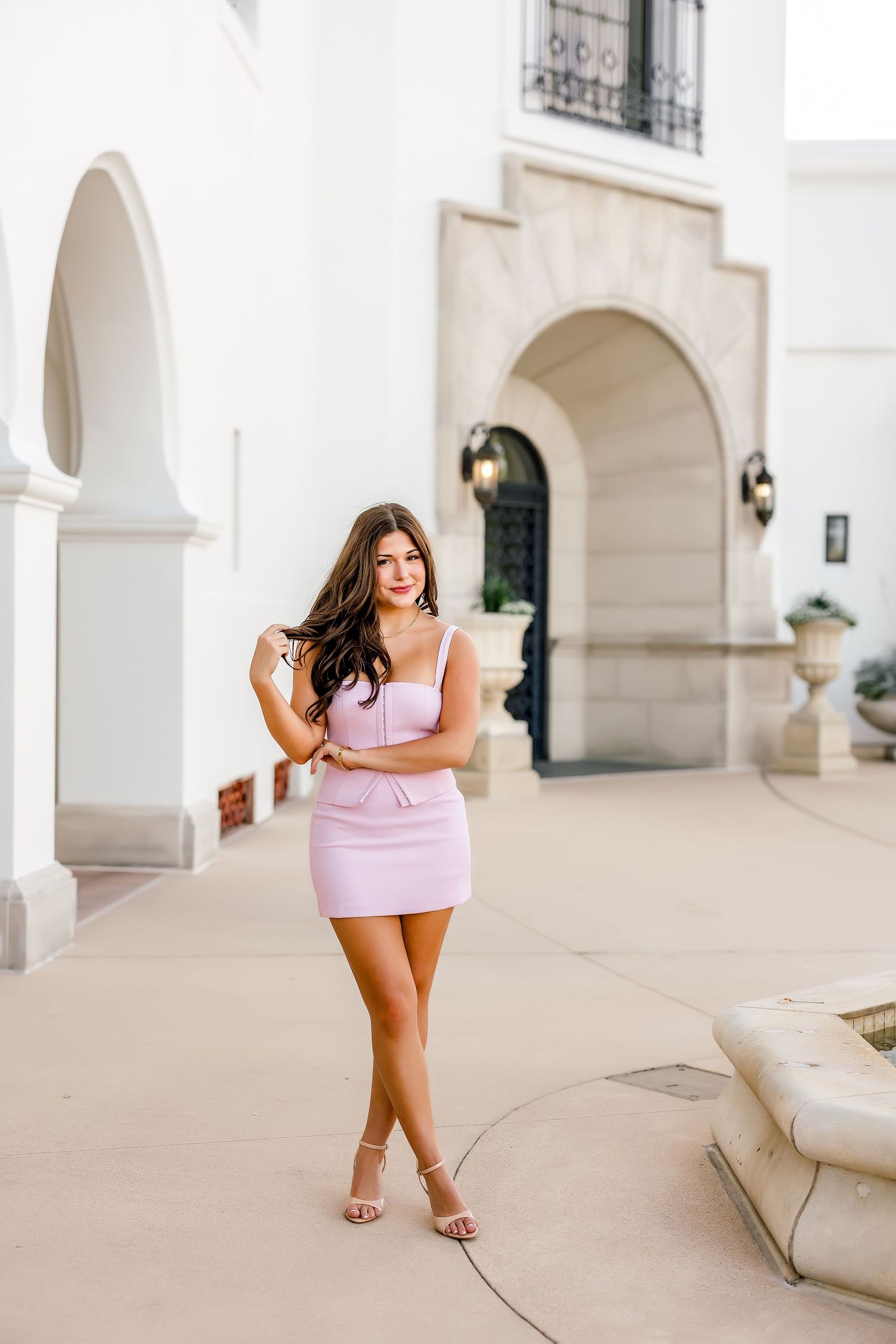 Young woman in pink mini dress and heels posing outdoors near white arched building architecture.