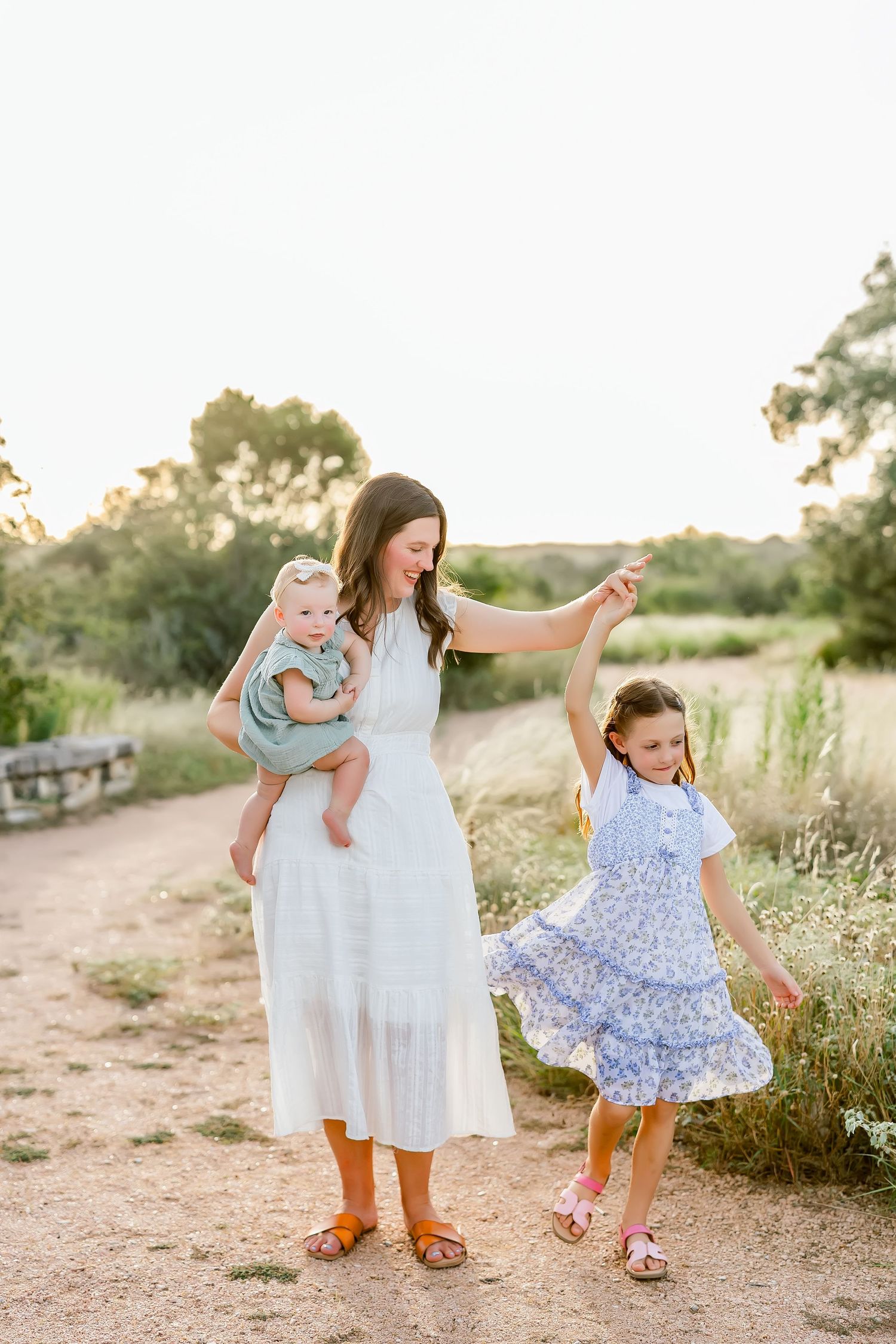 Mother in white dress holding baby while spinning young daughter in blue floral dress on sunny dirt path.