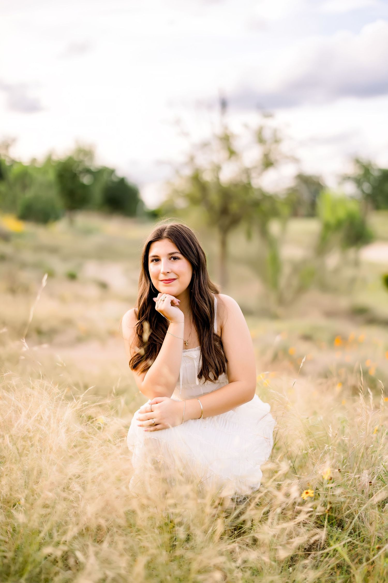 A brunette young woman in a white dress crouches gracefully in a golden field of tall grass.