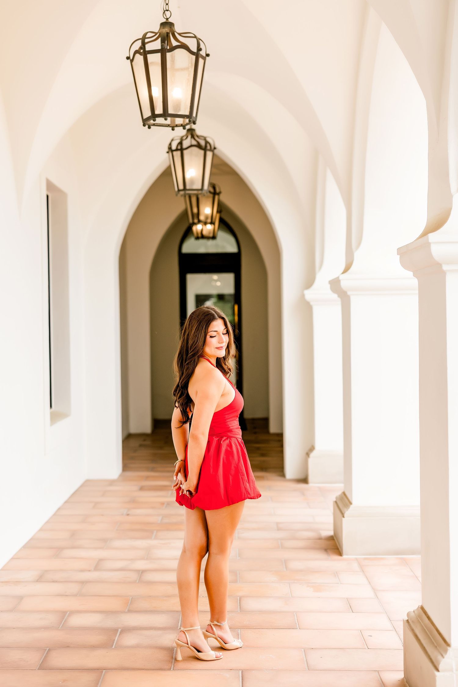 Young woman in red mini dress posing in elegant white arched hallway with hanging lanterns.
