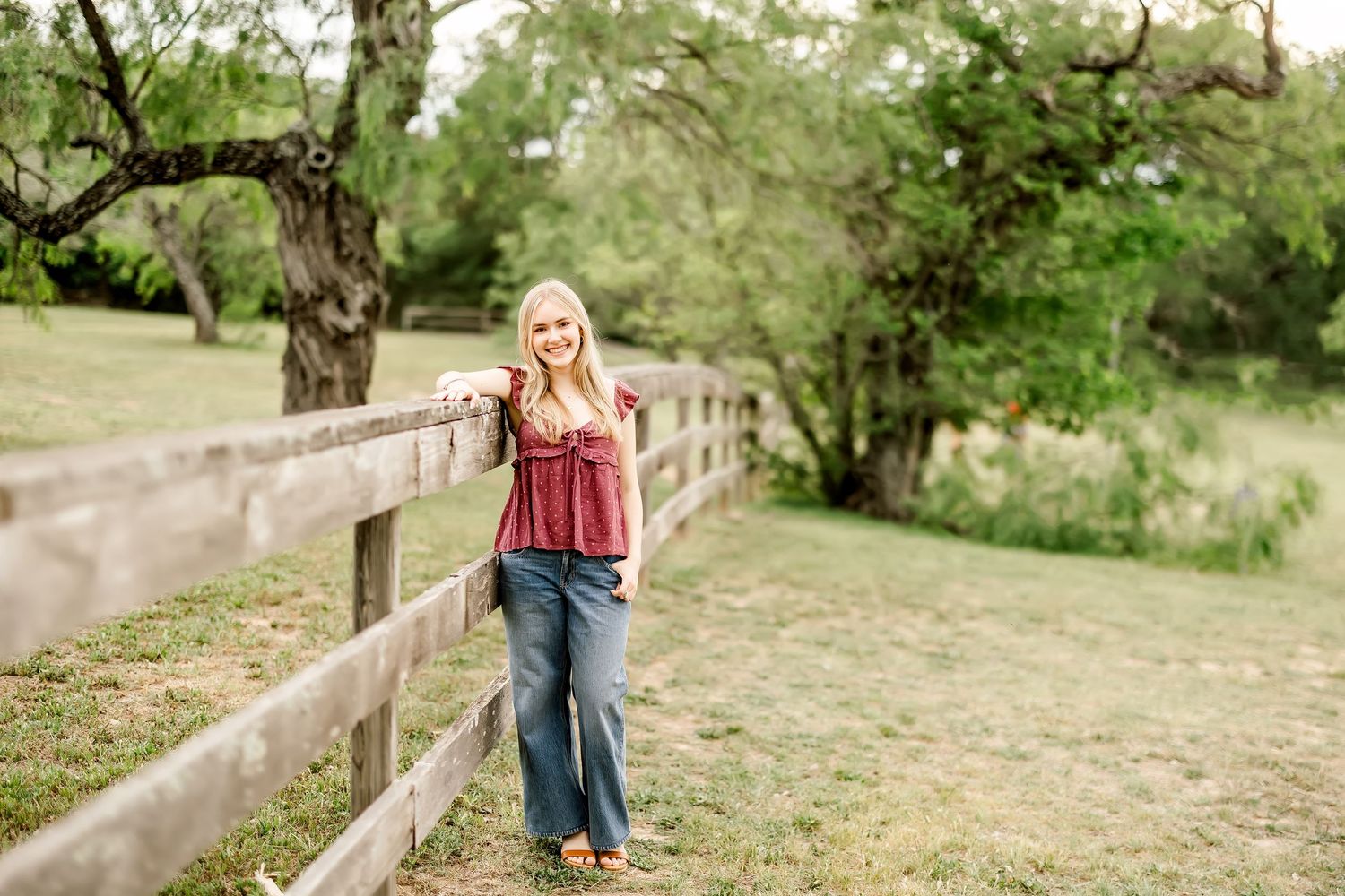 A blonde girl leans against a wooden fence in a green field  a senior portrait at a mini session in Commons Ford Ranch in Bee Cave, Tx.