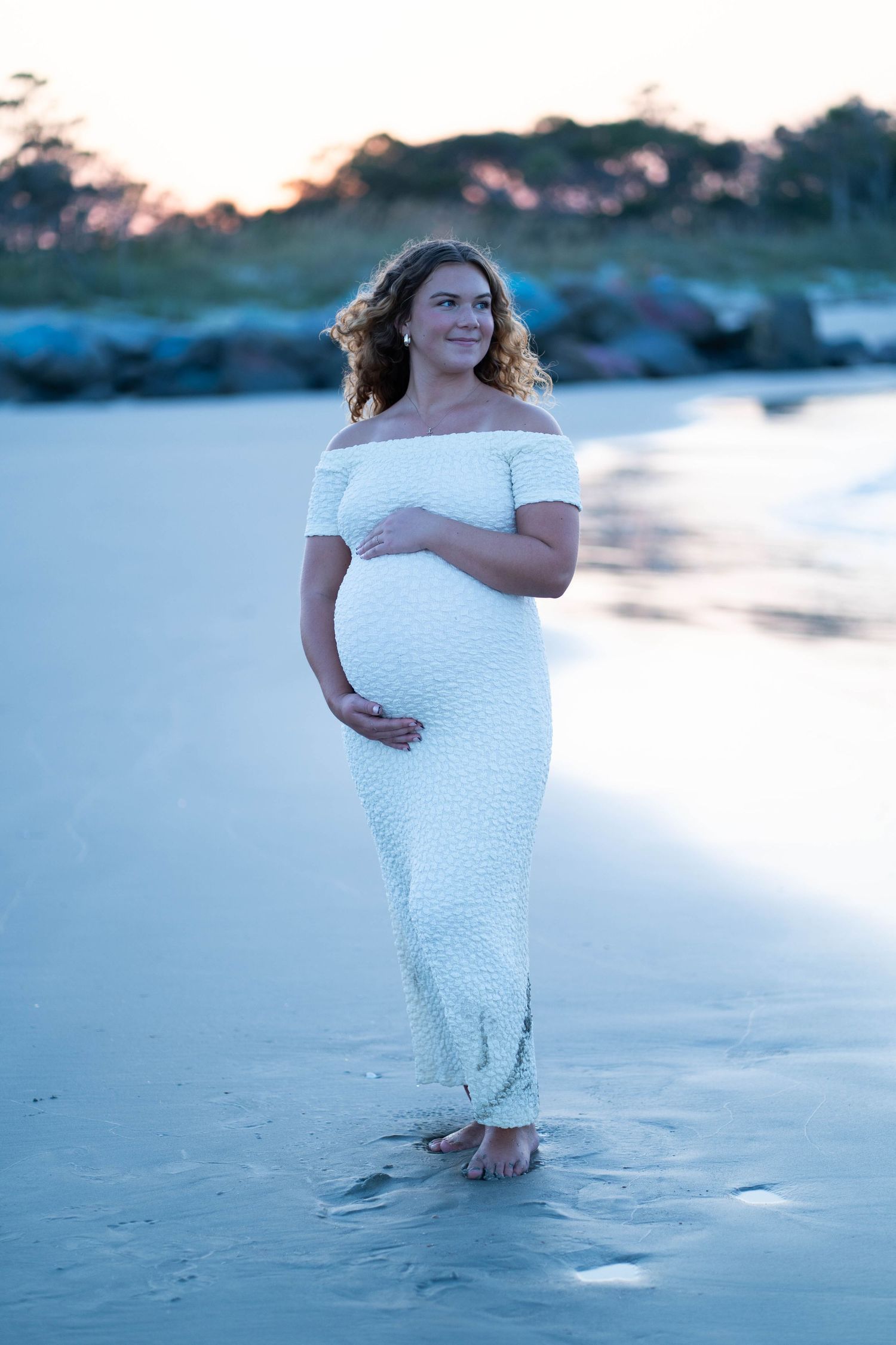 Woman in white dress standing on beach at sunset during maternity photoshoot.