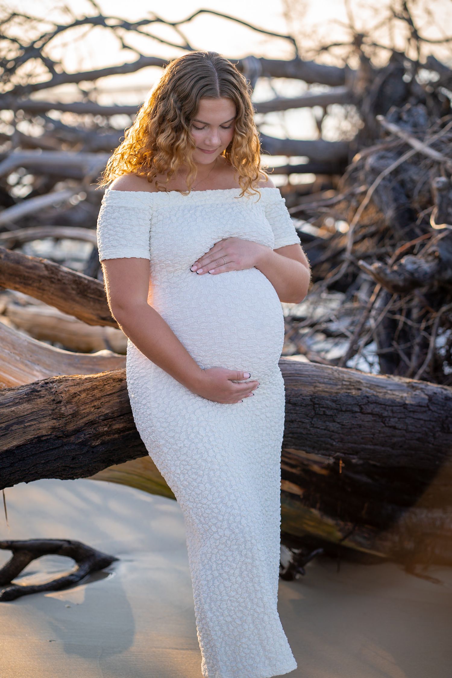 Maternity photoshoot on beach with driftwood, featuring woman in off-shoulder white dress at golden hour.