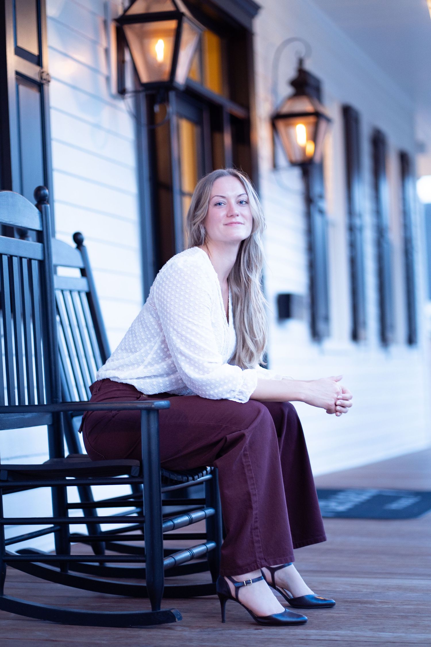 A person in a white sweater relaxes on a wooden rocking chair along a covered porch at dusk.