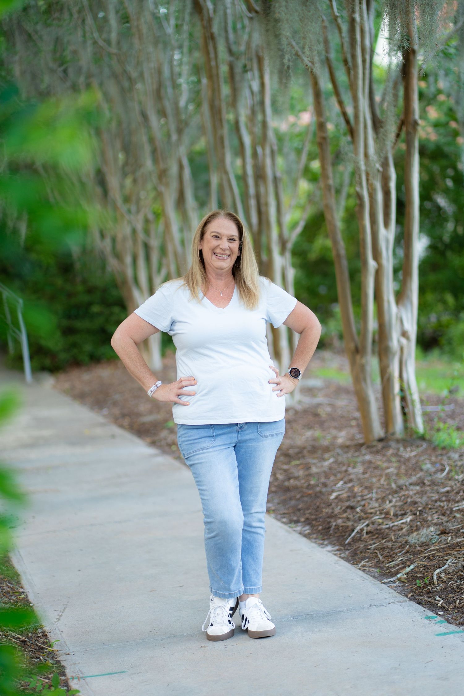 Someone in a white t-shirt and jeans stands on a paved path surrounded by tall birch trees in a park setting.