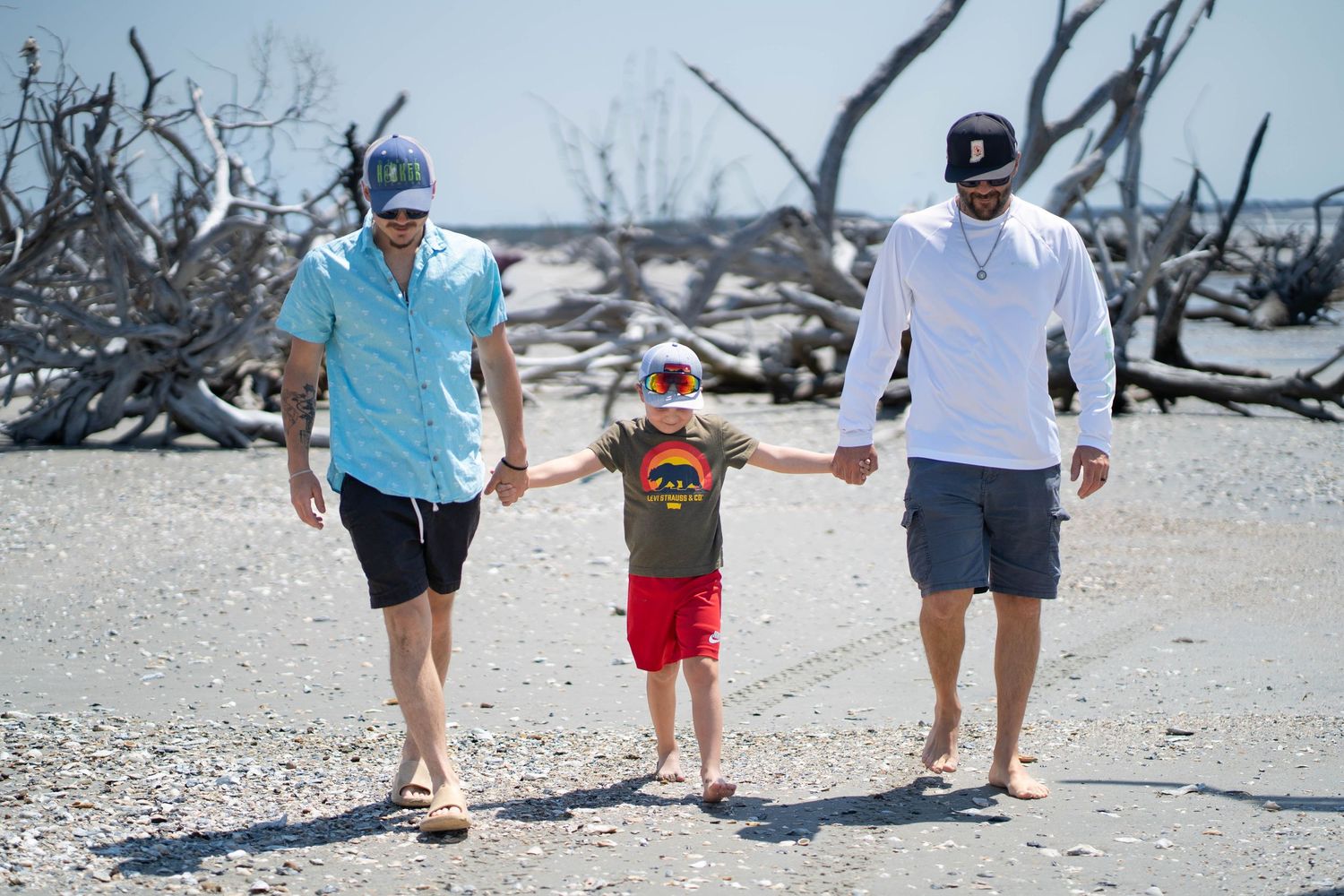 Family group walking together on sunny beach with driftwood in background.