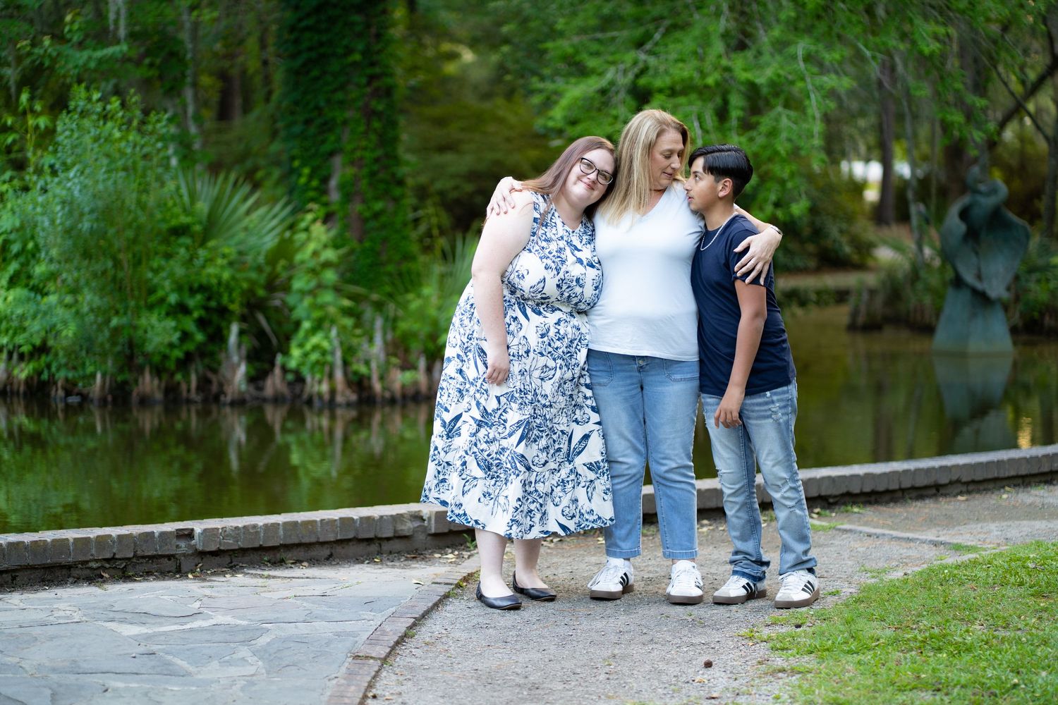 People walking dogs together on a paved trail surrounded by lush green trees next to a peaceful pond.