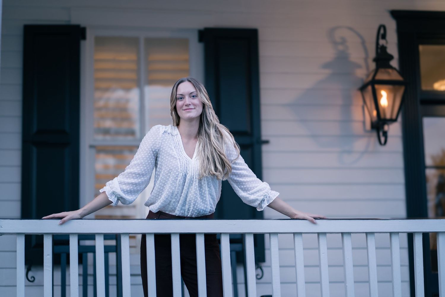 Woman in white blouse leans on white porch railing with black shutters in background.