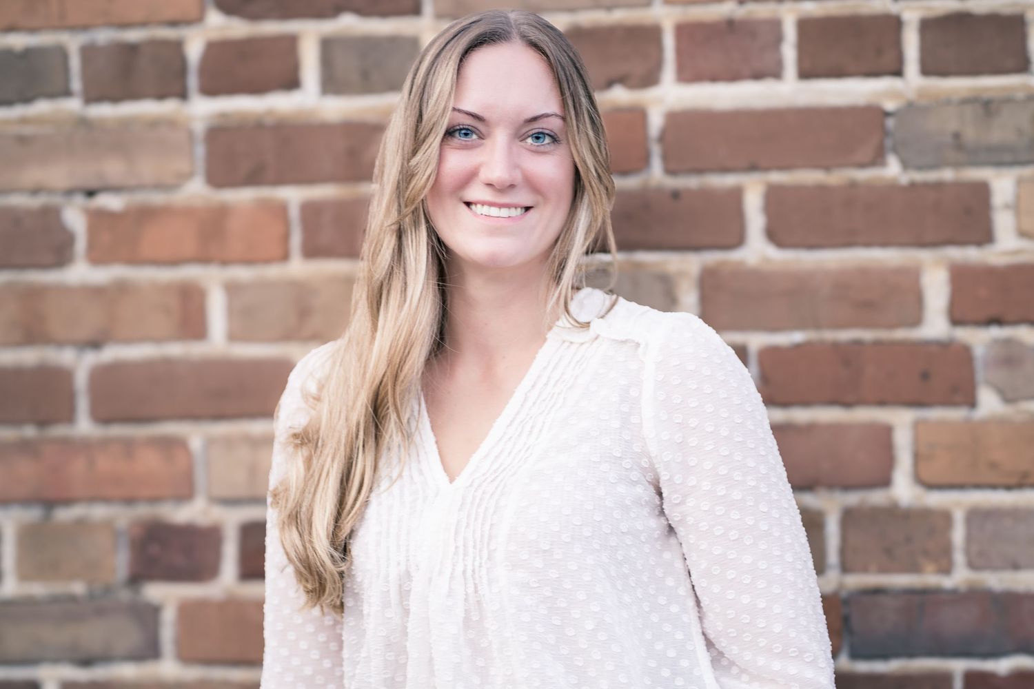 Portrait of woman in cream blouse smiling against brick wall background.