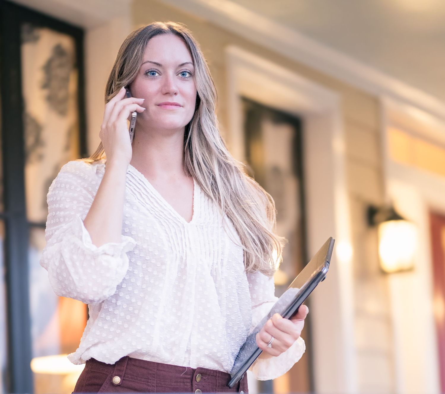 Young woman in a white ruffled top uses mobile phone while smiling in well-lit office hallway.