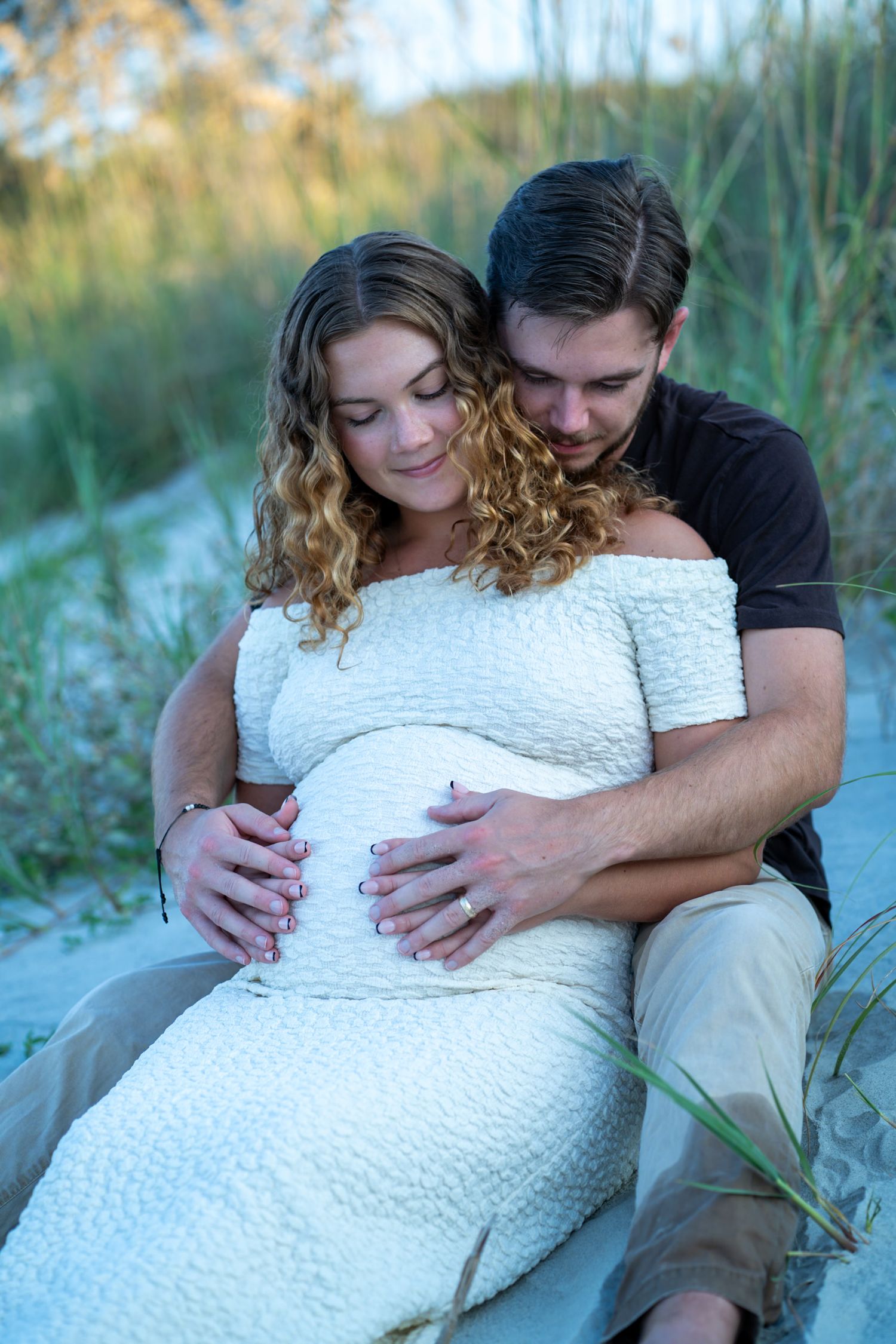 Maternity photo of couple embracing on beach with white lace dress during golden hour.