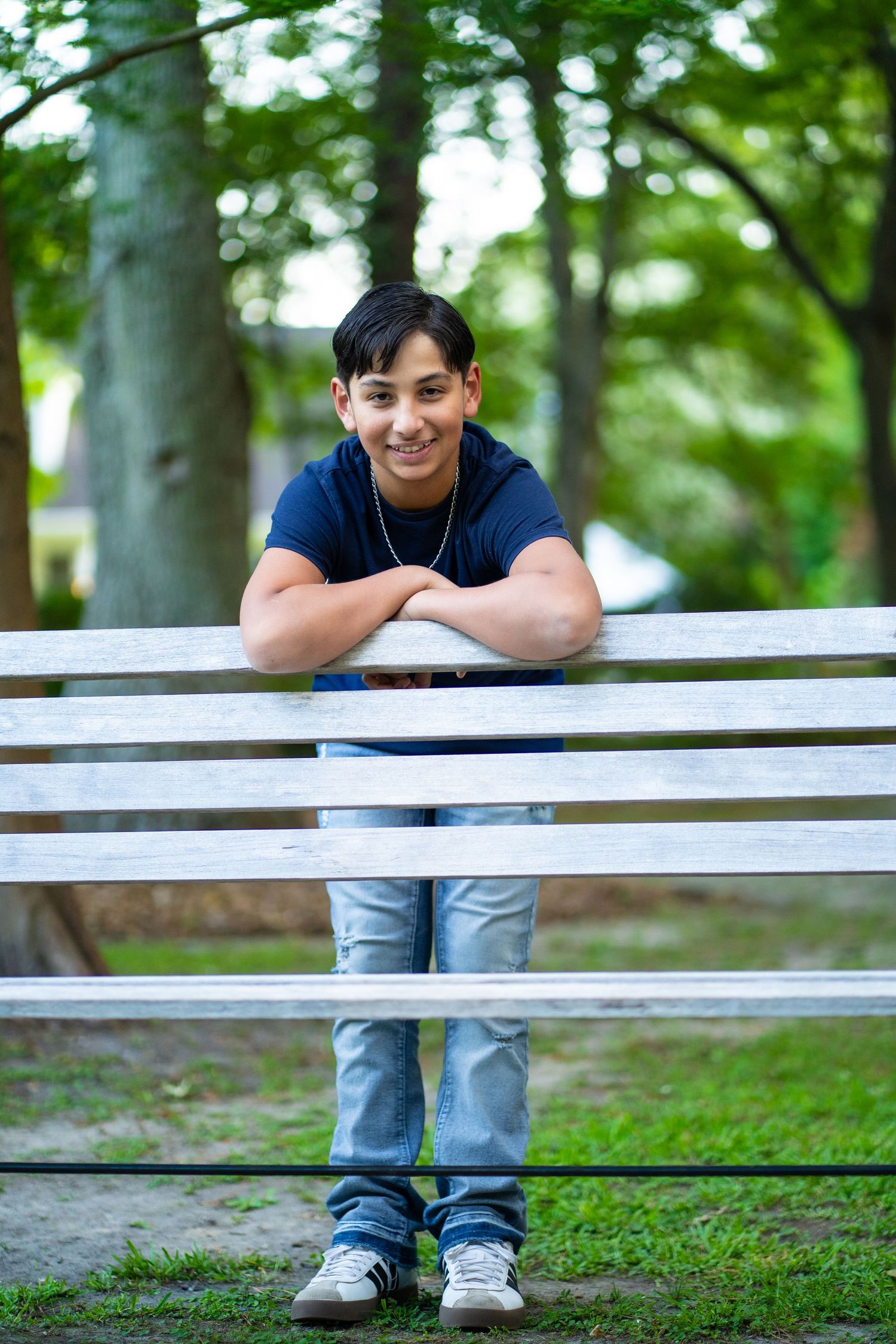 Person in blue jeans leans casually against white fence railing in wooded outdoor setting with green trees.