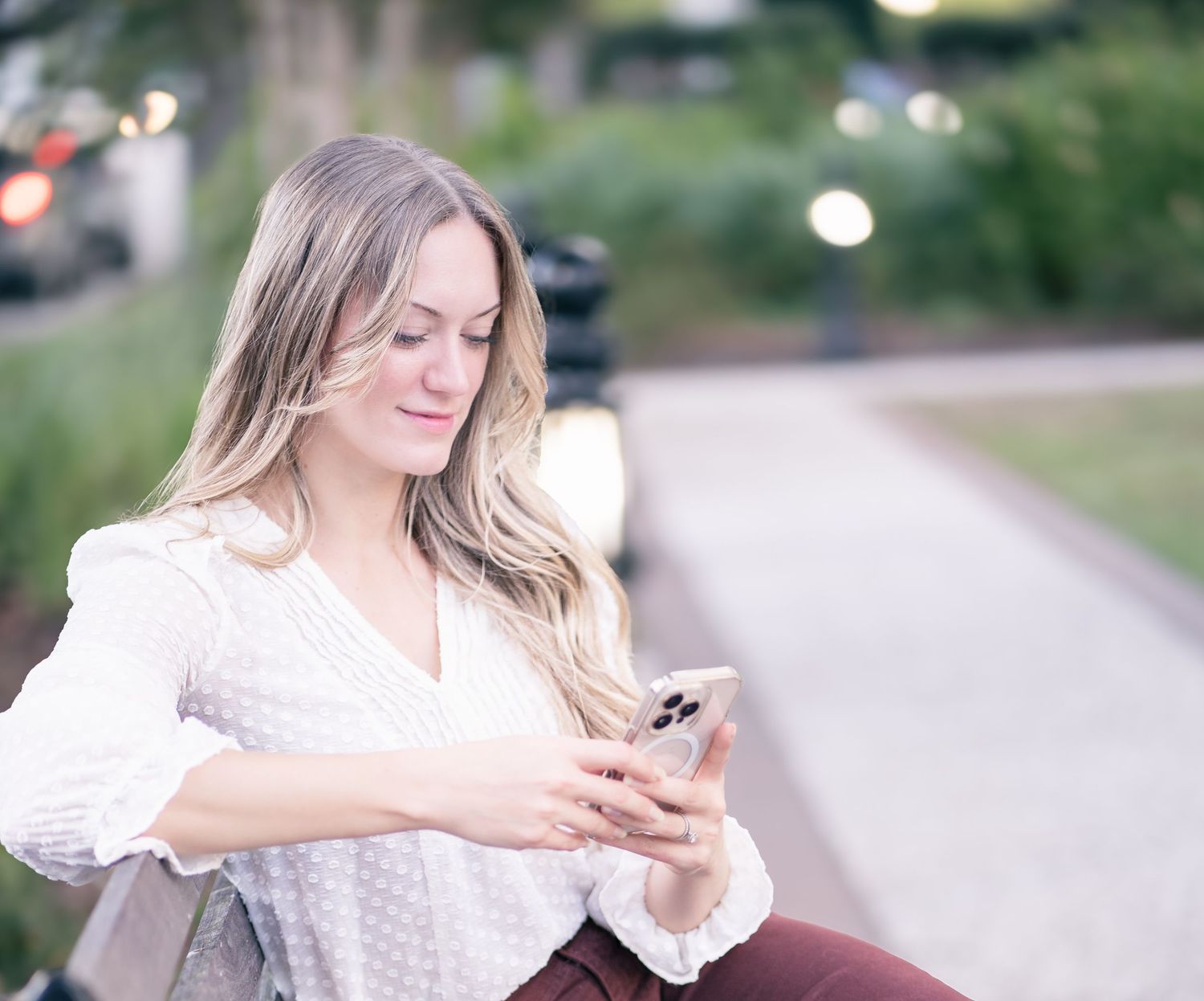Woman in white blouse checks phone while sitting outdoors on blurred background path.
