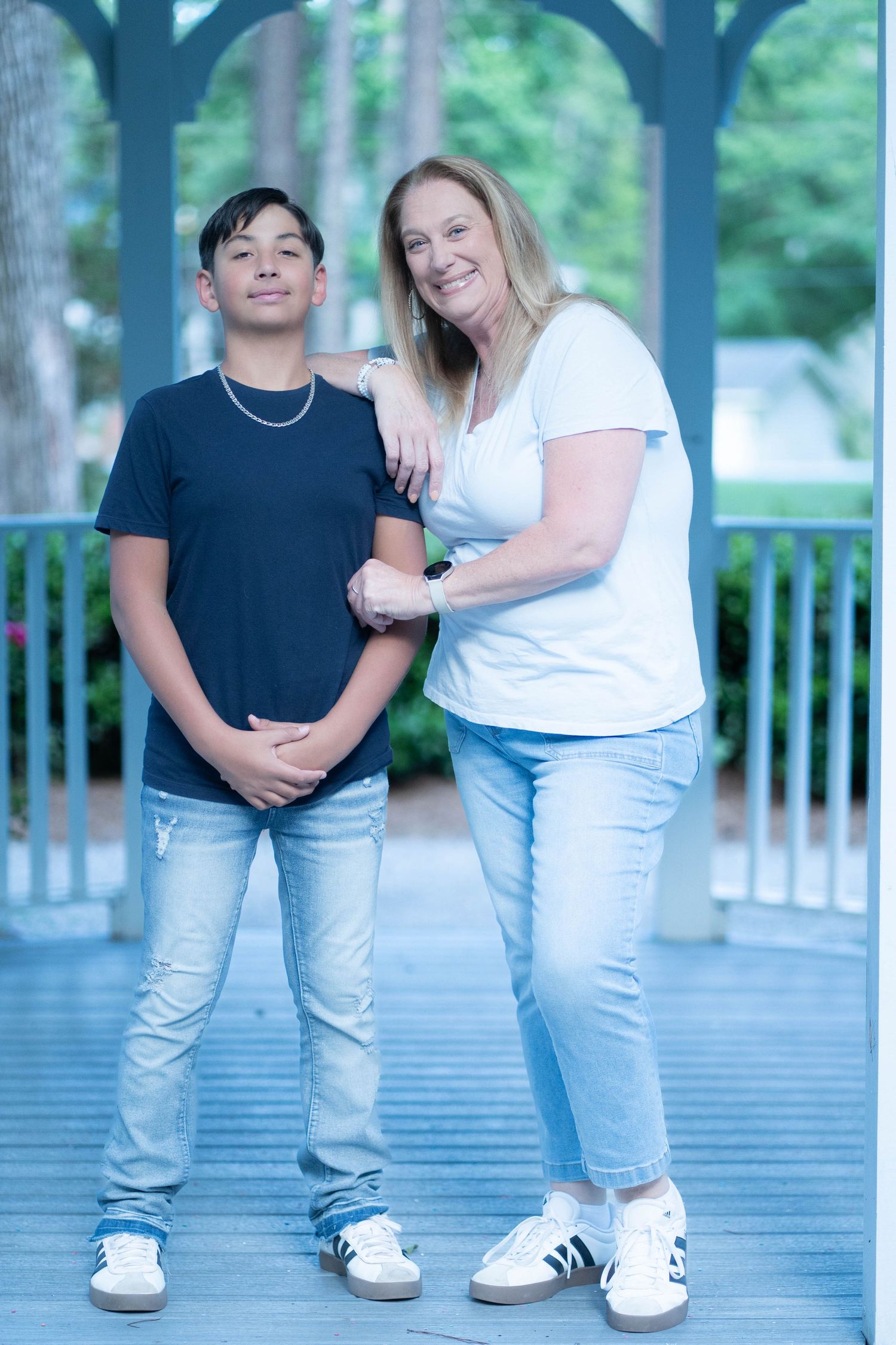 Two people posing together on a blue gazebo deck with green foliage behind.