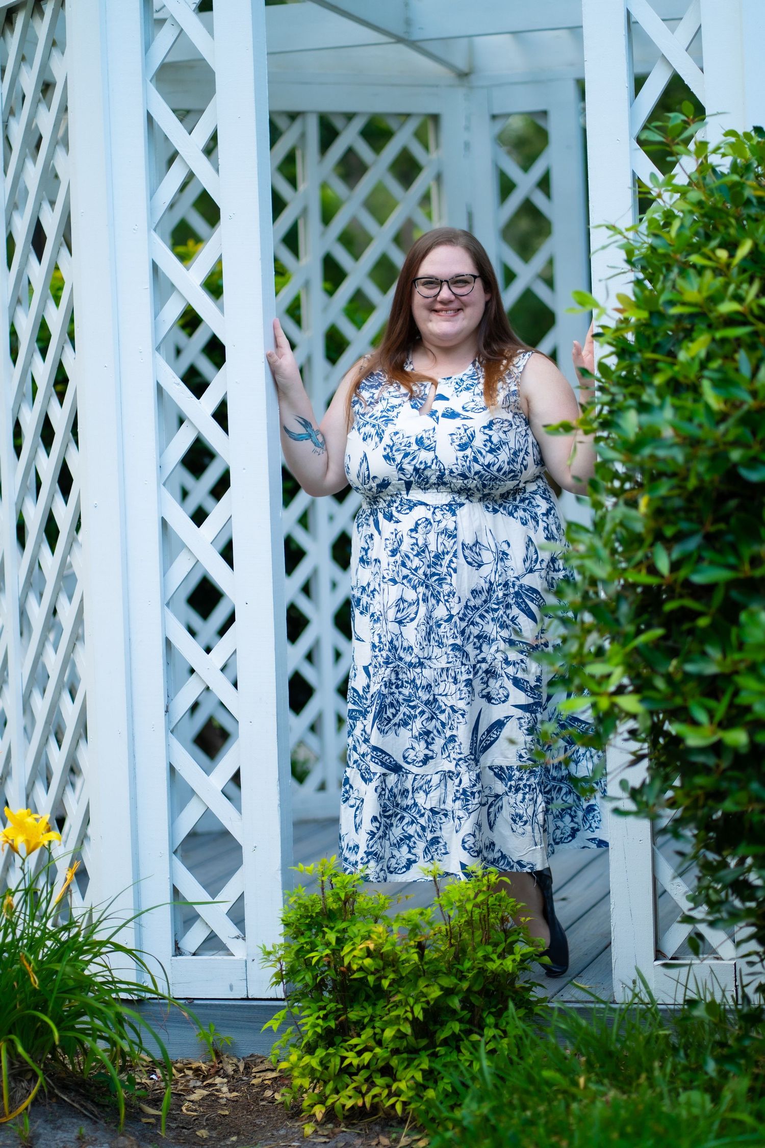 Woman in blue and white floral dress posing by white lattice garden trellis.